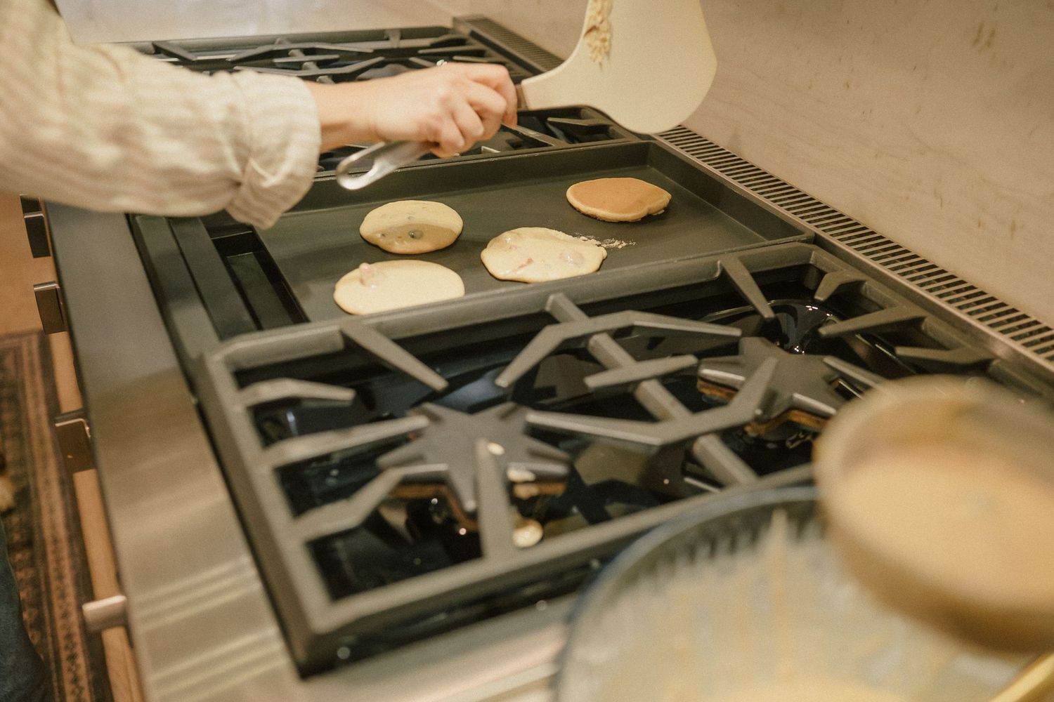 Close-up of pancakes cooking on a gas stove griddle, with a hand holding a spatula nearby.
