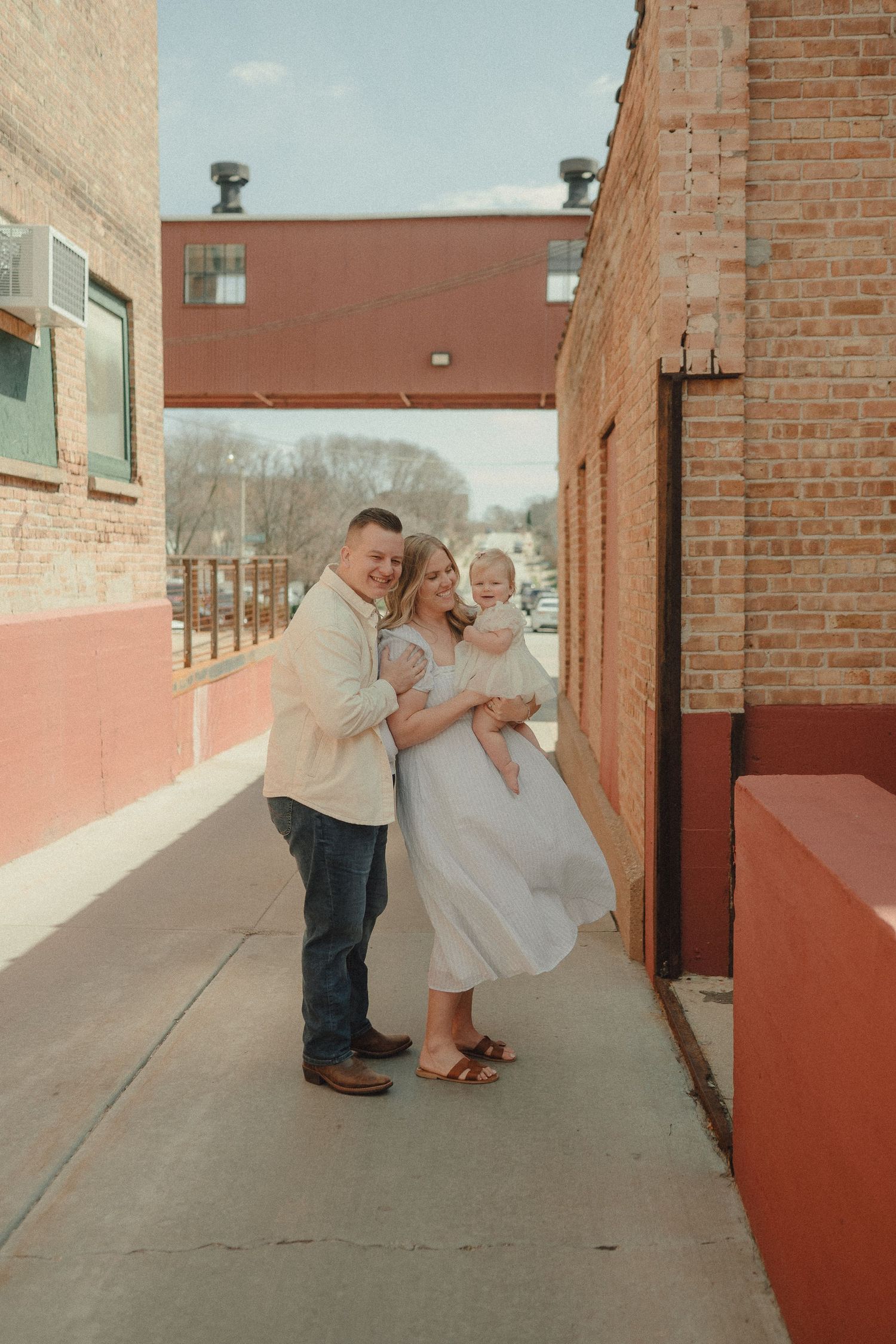 Young family with baby poses on urban sidewalk between brick buildings for a professional photo session.