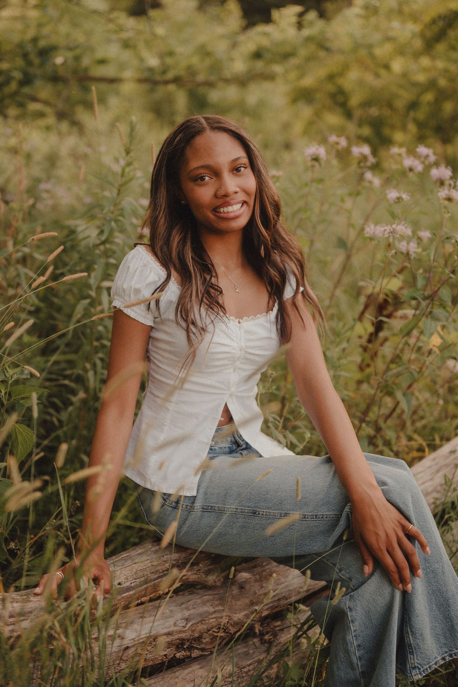 Young woman smiling in white top and jeans, sitting on a log surrounded by wildflowers in a lush green outdoor setting.