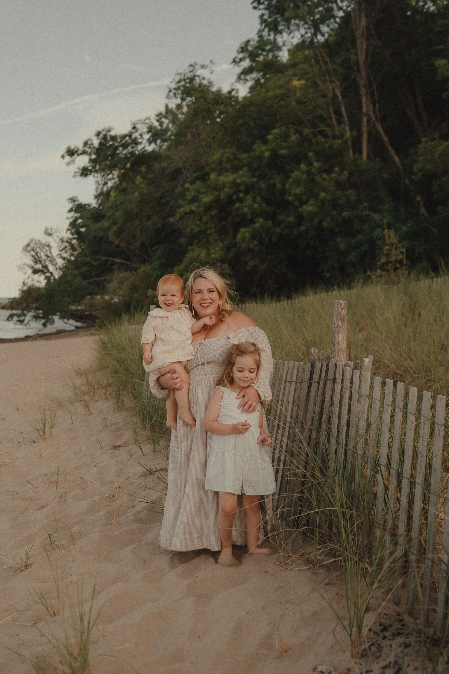Smiling mother holding baby while daughter hugs her on sandy beach.