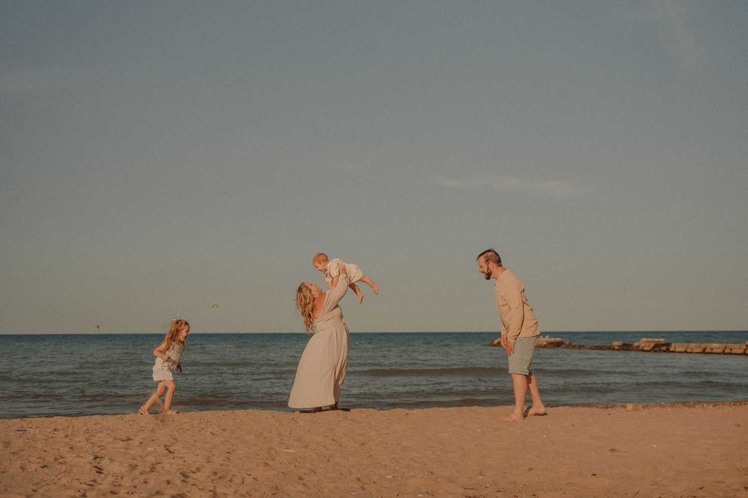 Family of four enjoying a sunny beach day, with parents lifting a baby while a young girl plays on the sand near the ocean.