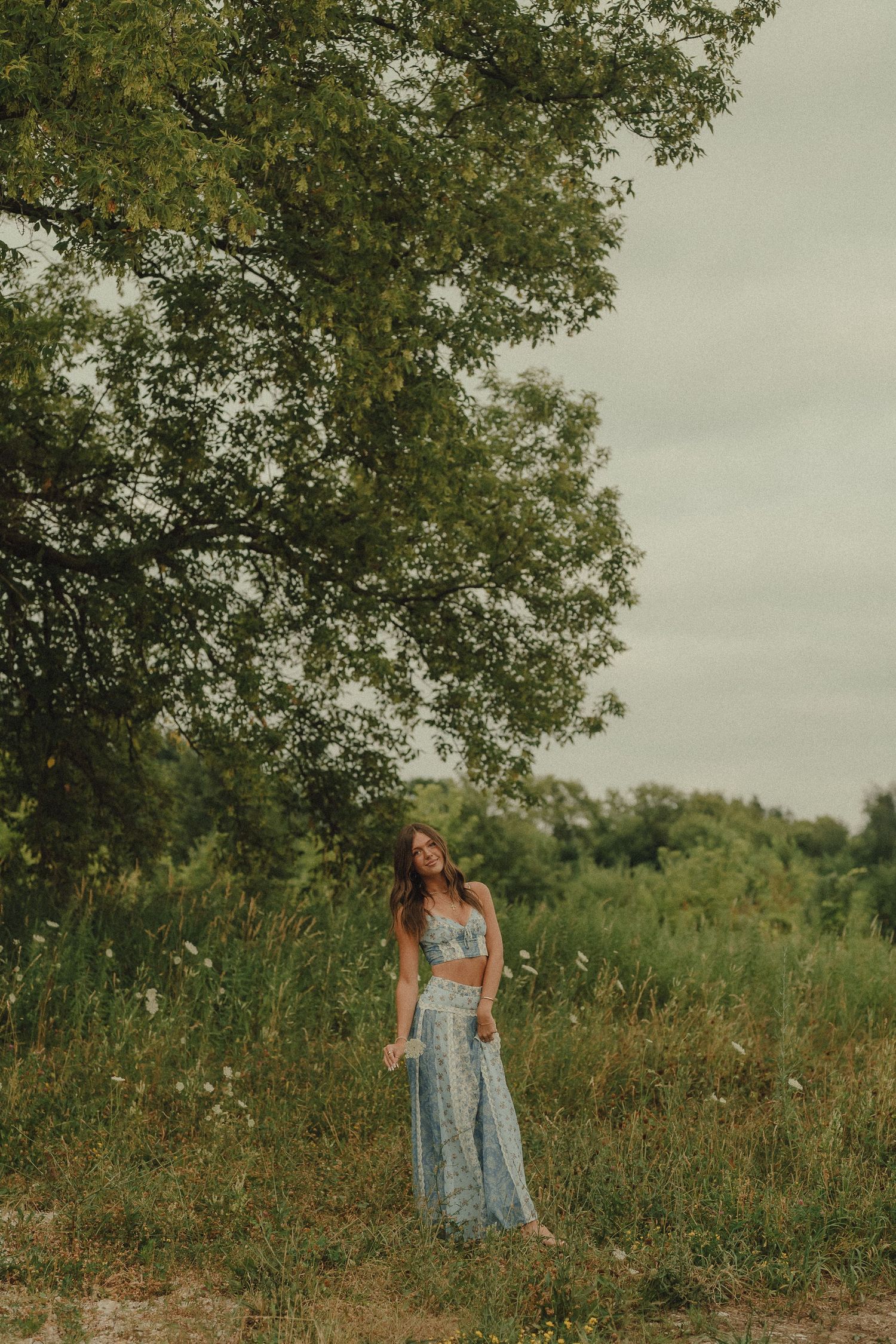 Young woman in blue floral two-piece outfit standing in a lush green field beneath a large tree.