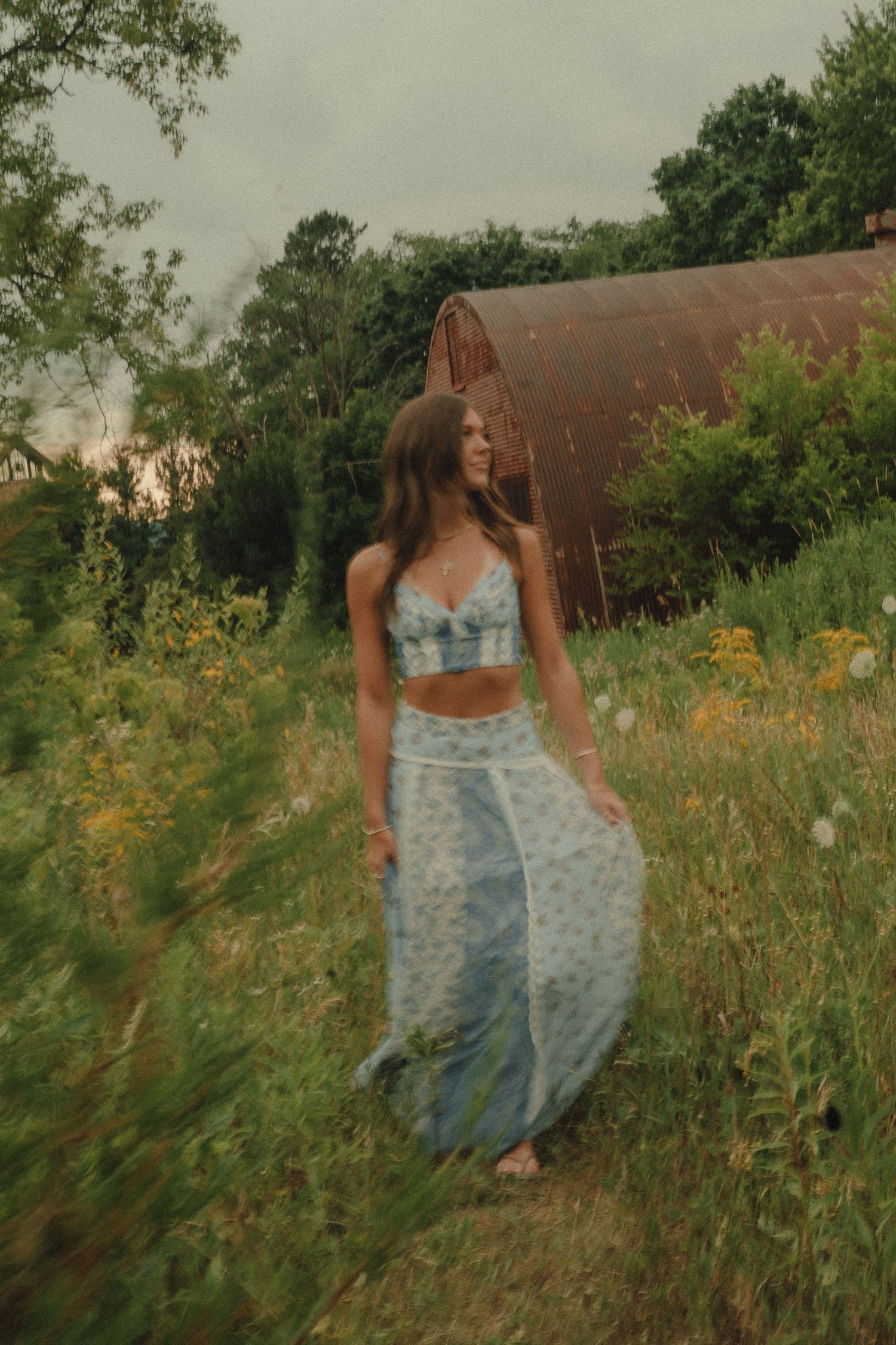 Woman in blue floral crop top and maxi skirt walking through green meadow with vintage barn backdrop.