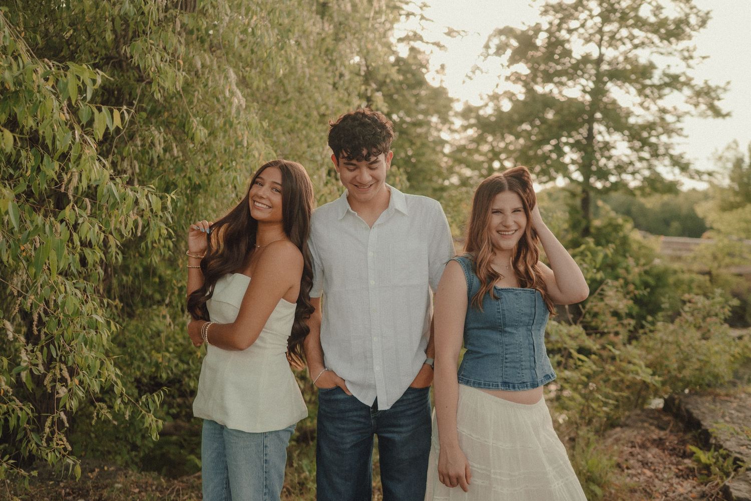 Three young adults smiling and posing together outdoors among lush green trees on a warm sunny day.