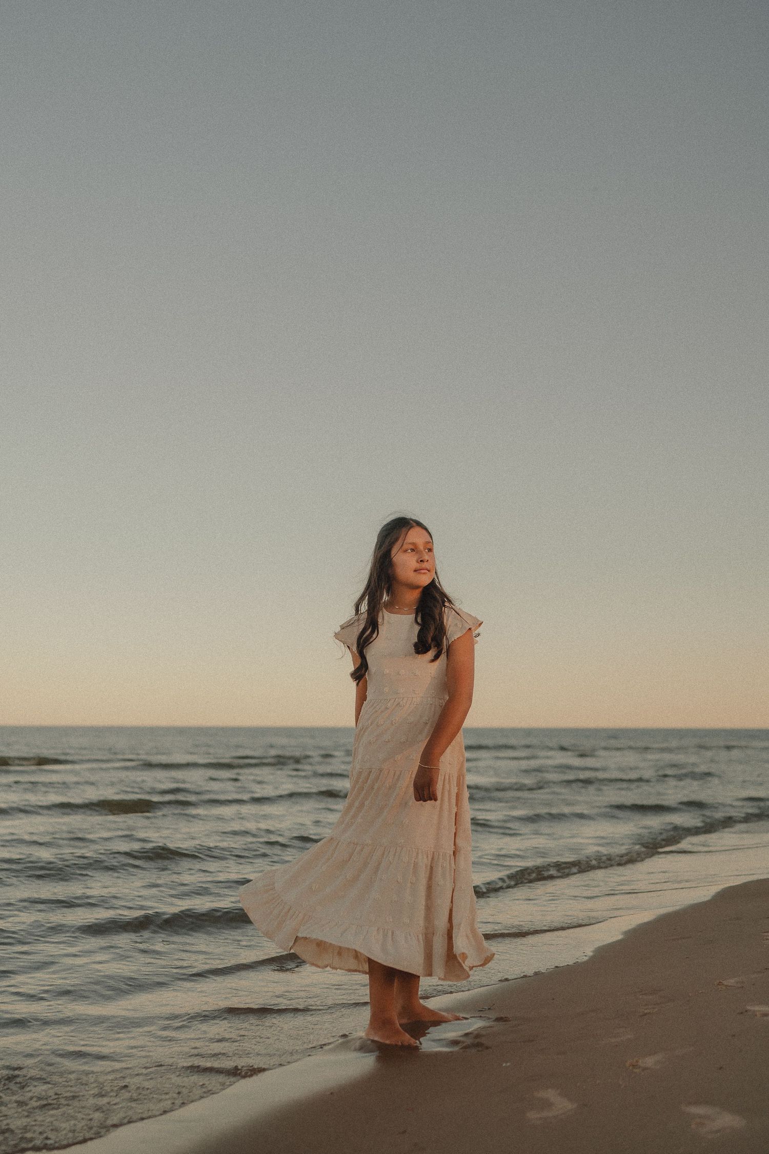 Young woman in white dress standing barefoot on beach shore at golden hour sunset.