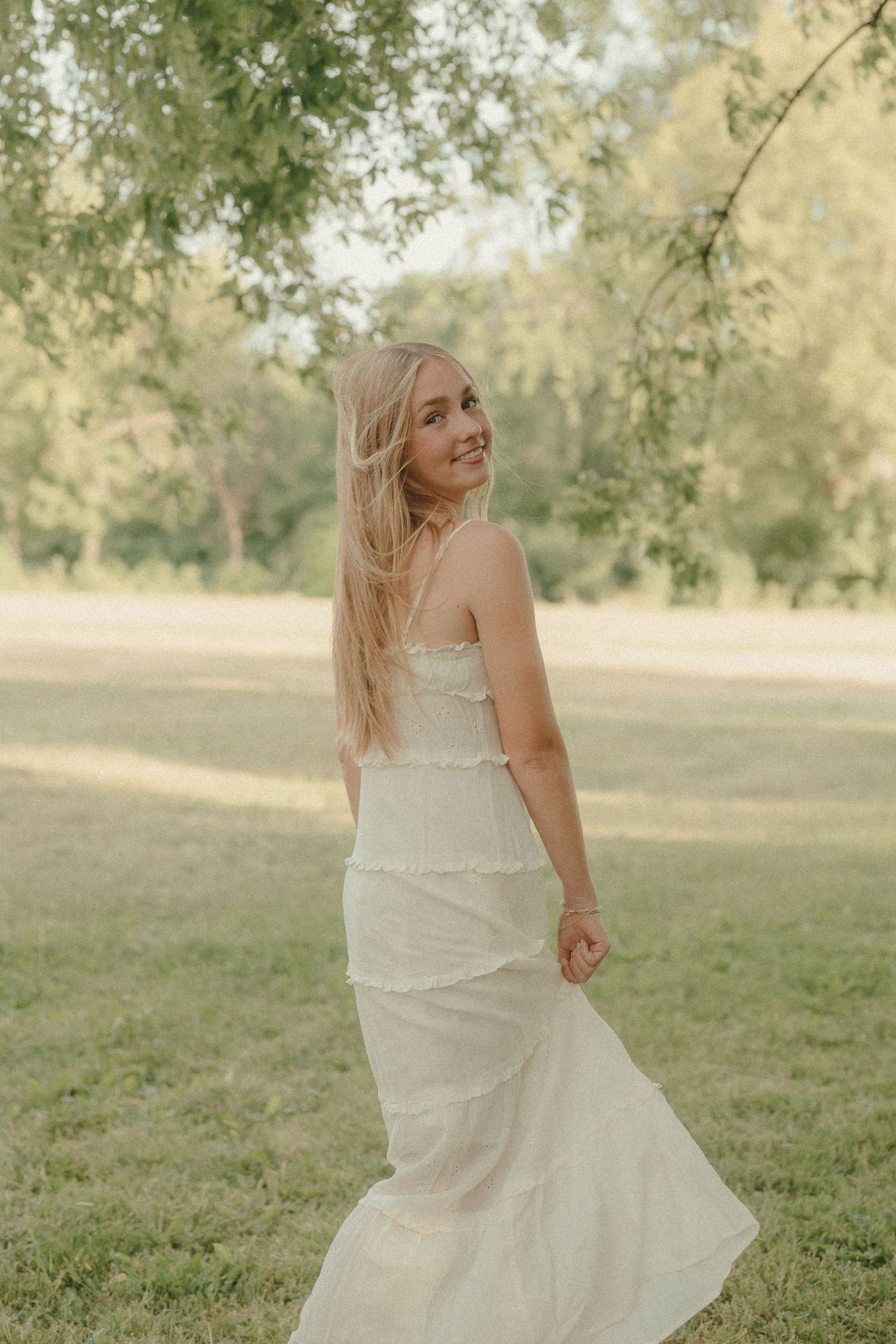 Young blonde woman in white maxi dress smiling over shoulder in sunlit green park setting.