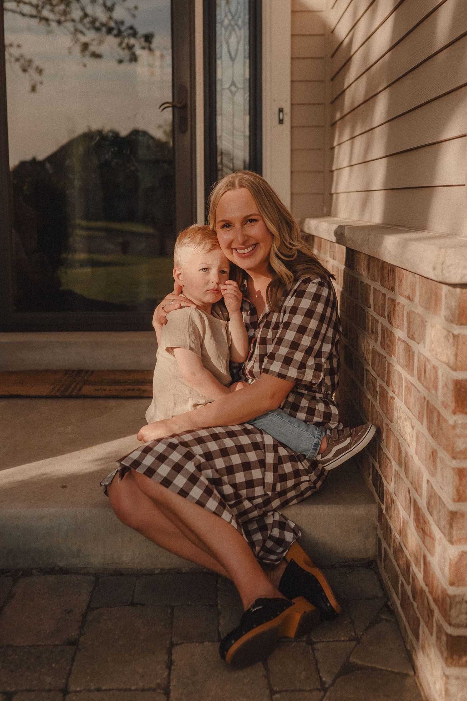 A smiling blonde mother in a plaid dress sits on porch steps cuddling her young toddler in warm light.