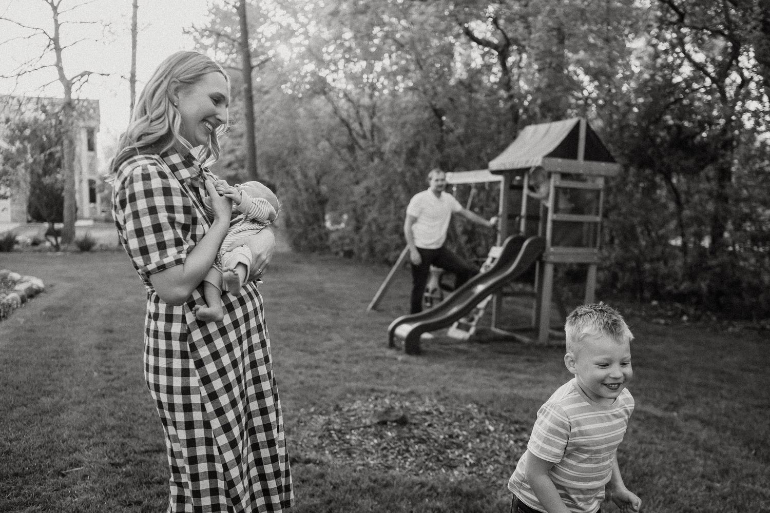 Black and white photo of a smiling mother holding a newborn as a laughing toddler runs nearby in a green backyard.