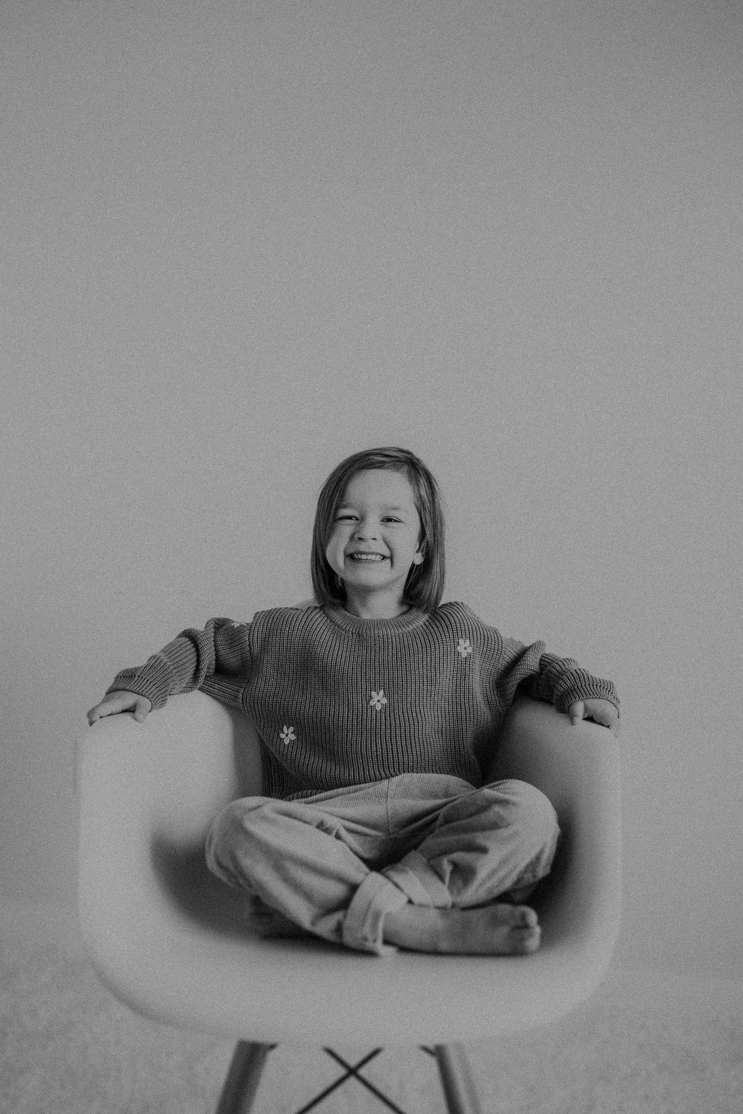 Laughing child relaxes in modern chair wearing cozy sweater in black and white photo.