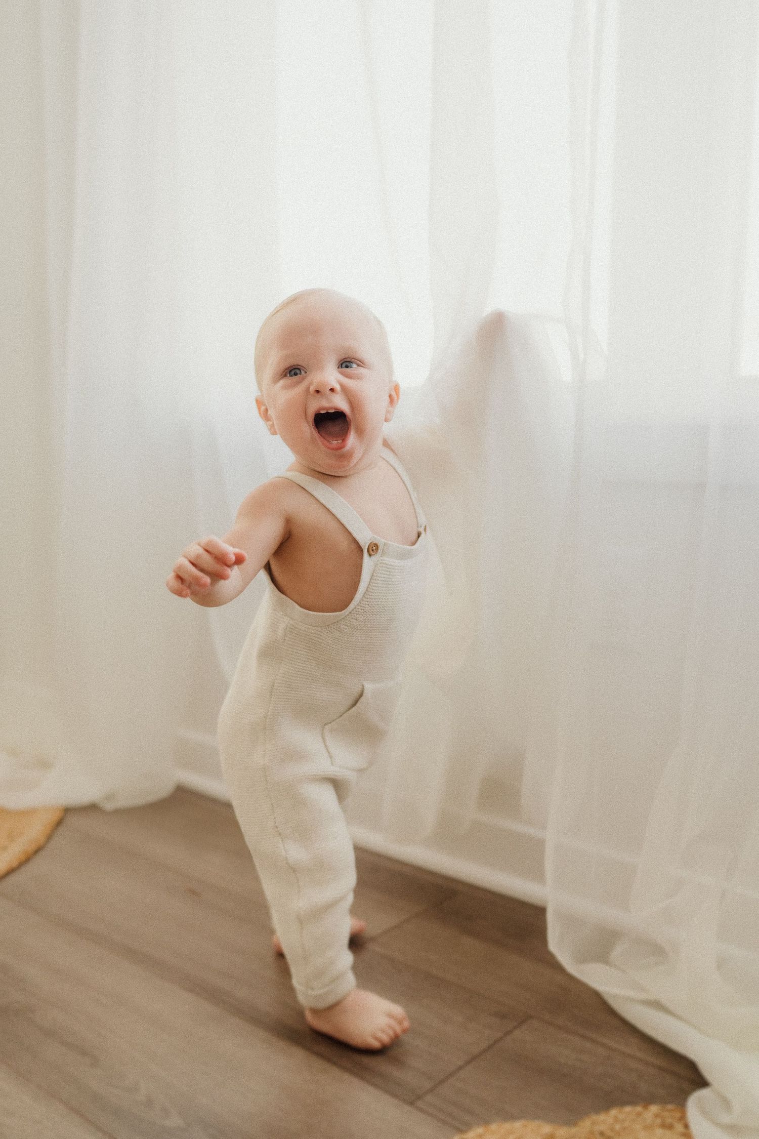 Happy baby in cream-colored romper standing independently by sheer white curtains.