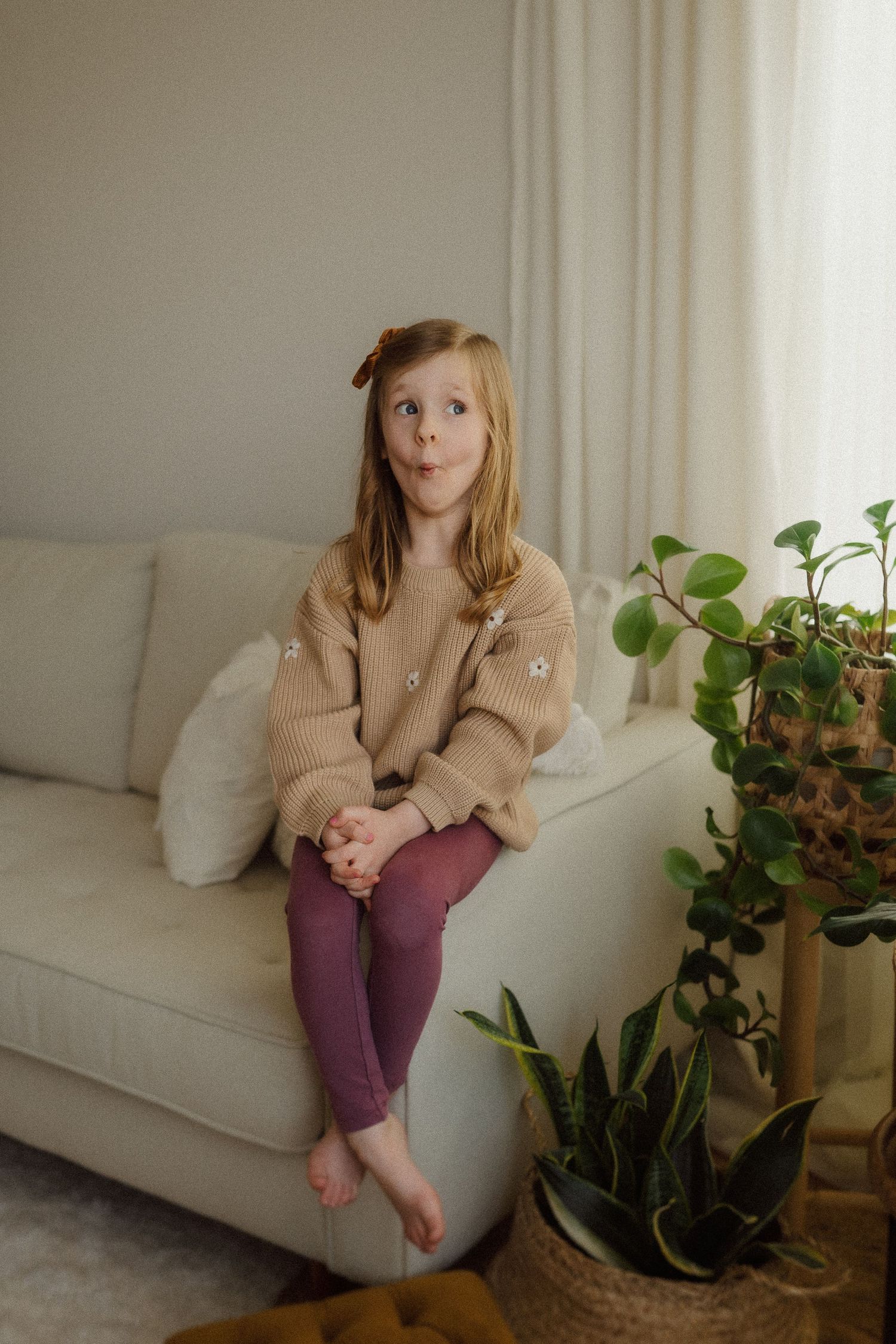 Child in beige sweater and purple leggings sits casually on white couch next to potted plants in cozy living room setting.