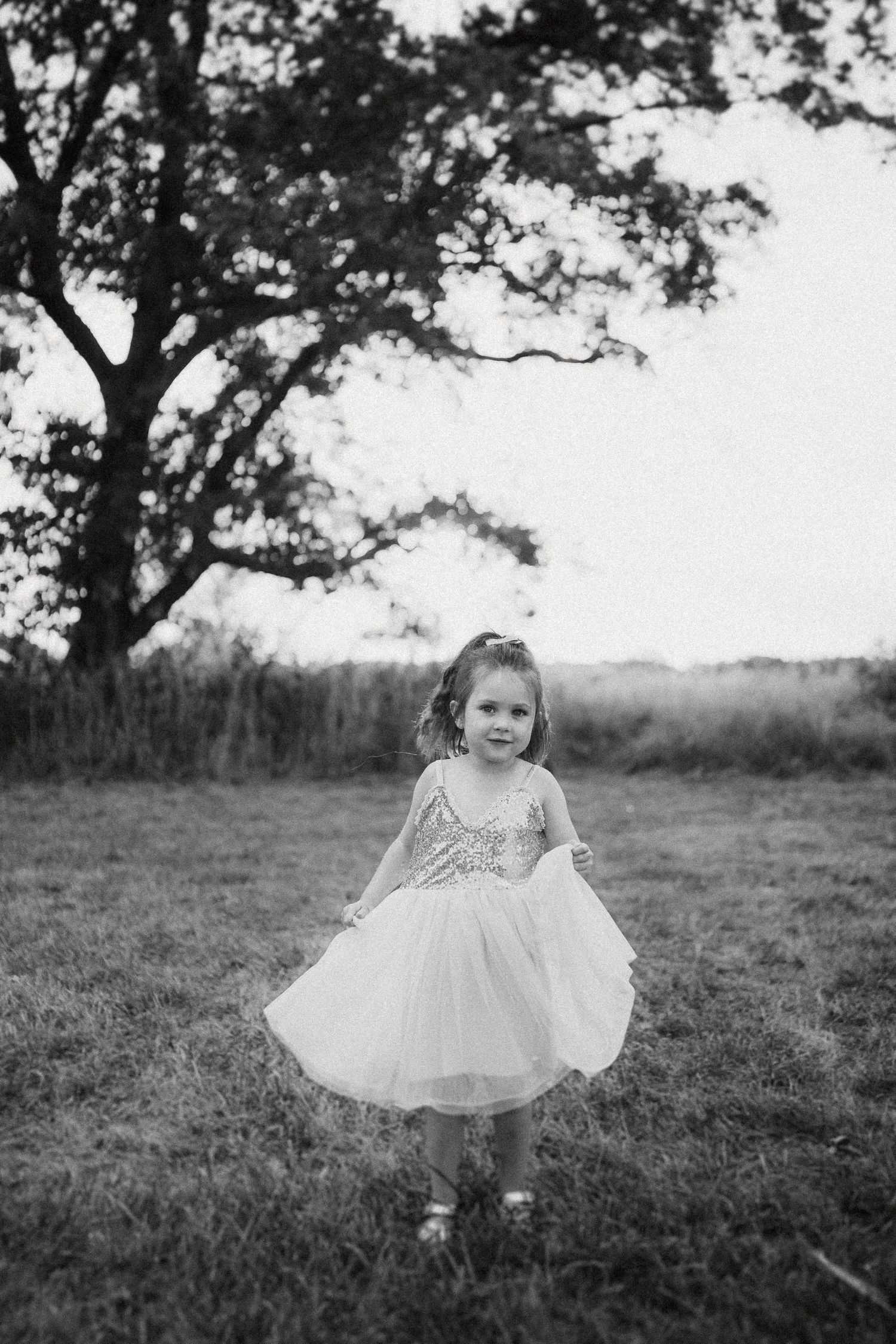 A small figure in a white party dress stands in a field with a large tree silhouetted in the background.