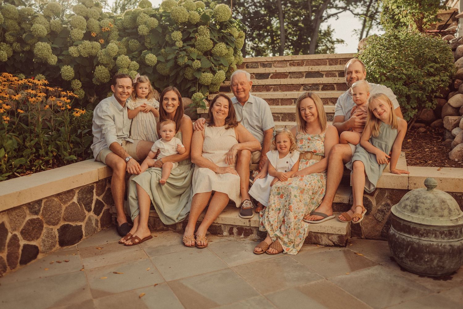 A family group poses together on rustic stone steps with hydrangea bushes in warm sunset lighting.