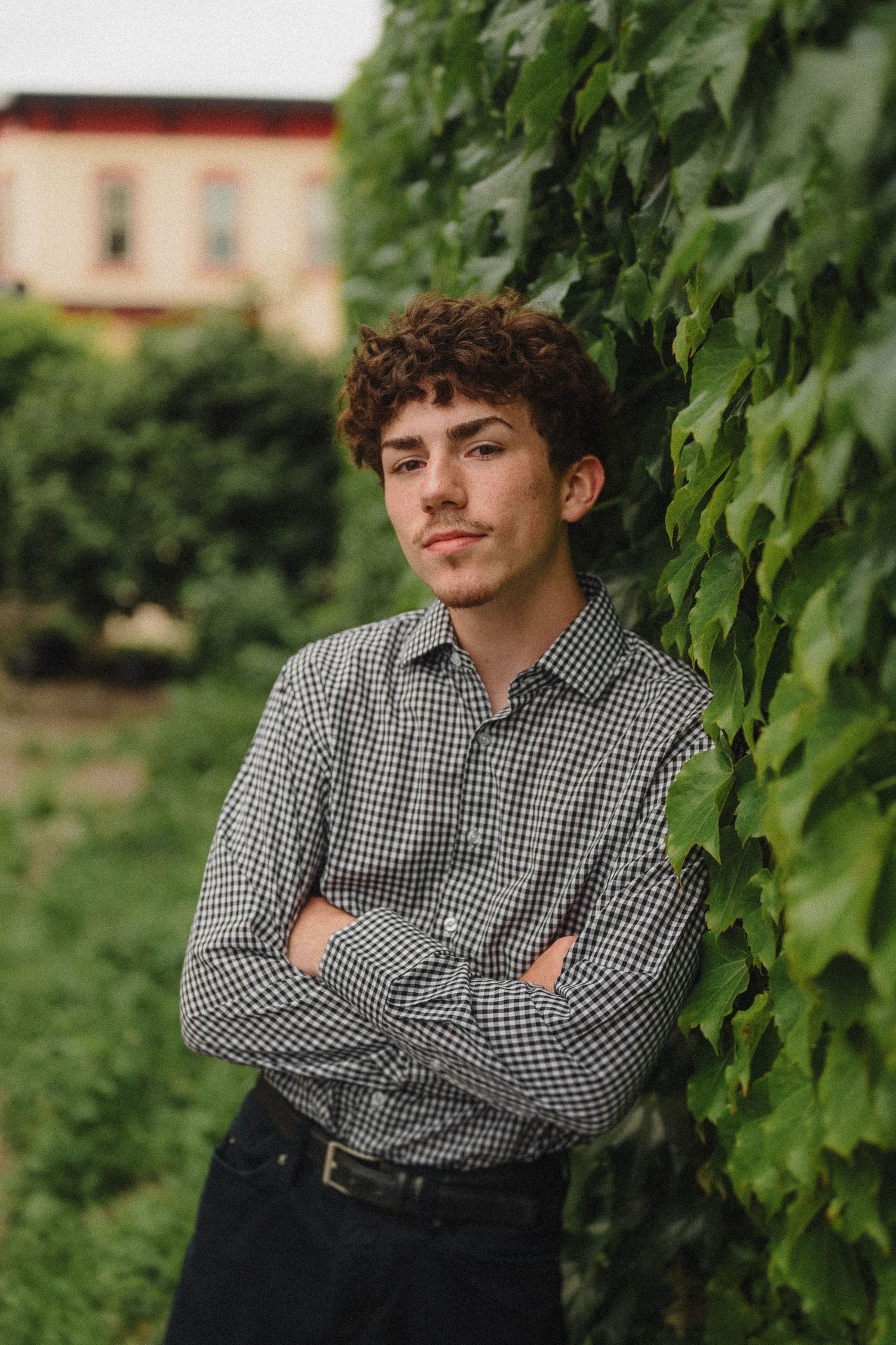Relaxed portrait of someone in a checkered shirt leaning against a vine-covered wall with greenery in the background.