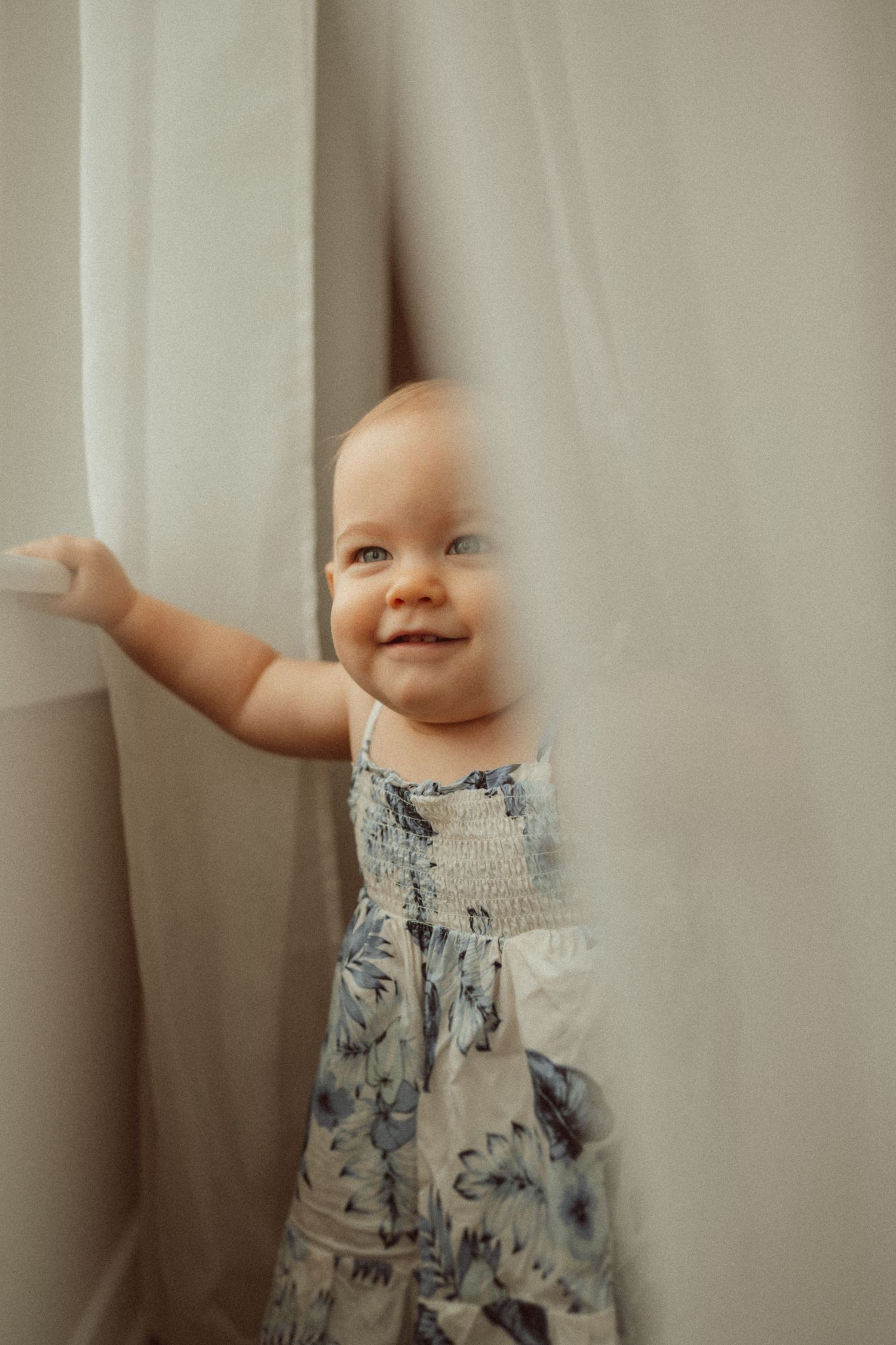 Baby wearing floral dress reaches up to touch white curtains while smiling.