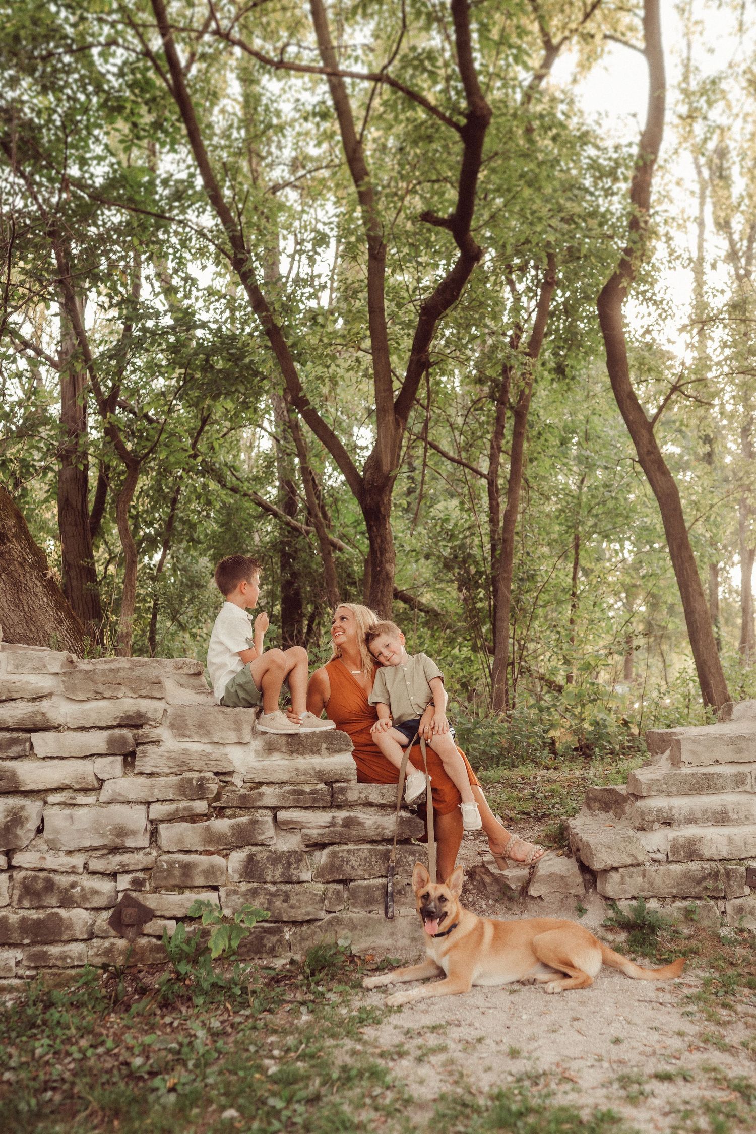 Two dogs join a family relaxing on ancient stone steps in a wooded setting during golden hour.