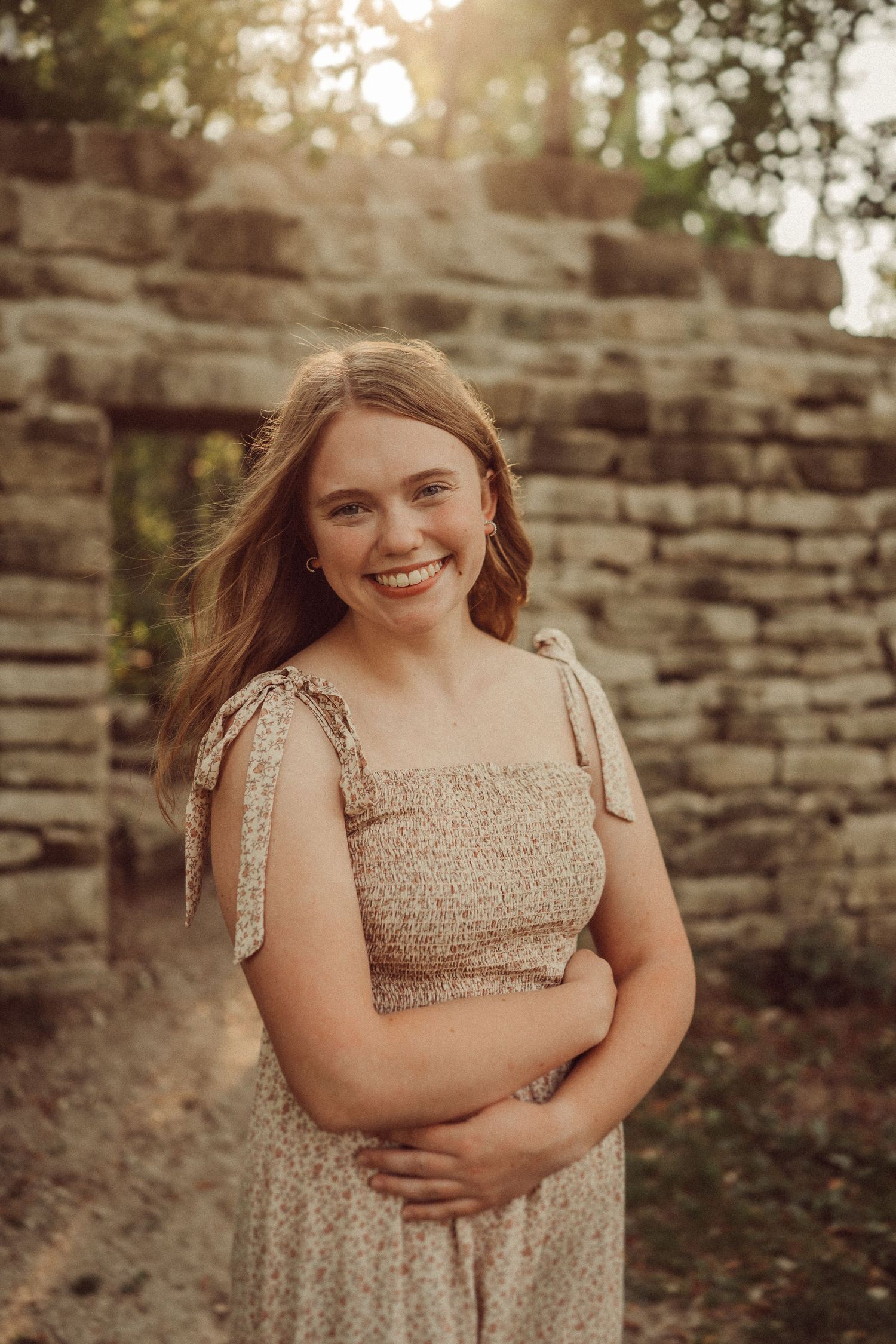 Portrait of a joyful woman in a beige summer dress standing by a stone wall at sunset.