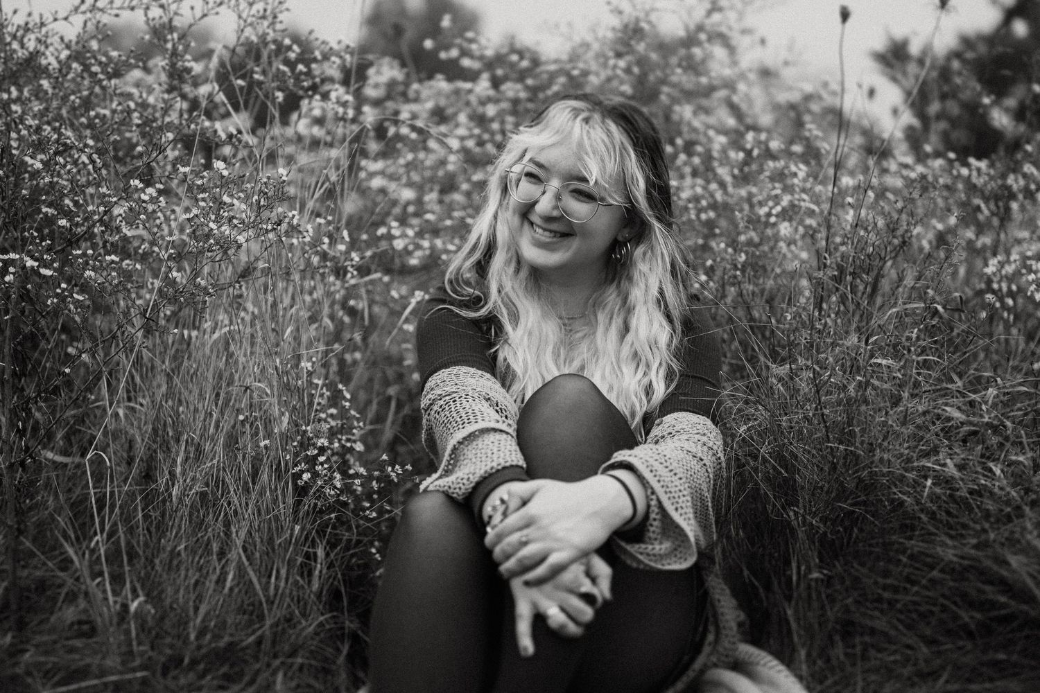 A joyful candid black and white portrait of someone sitting in a wildflower field wearing glasses.