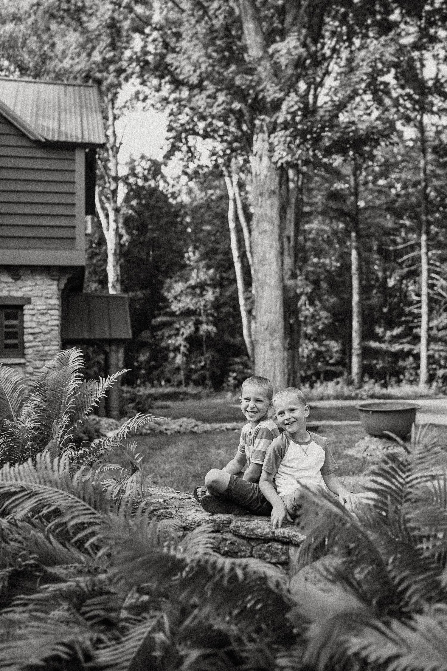 Children sitting on stone wall surrounded by lush ferns near rustic cabin.