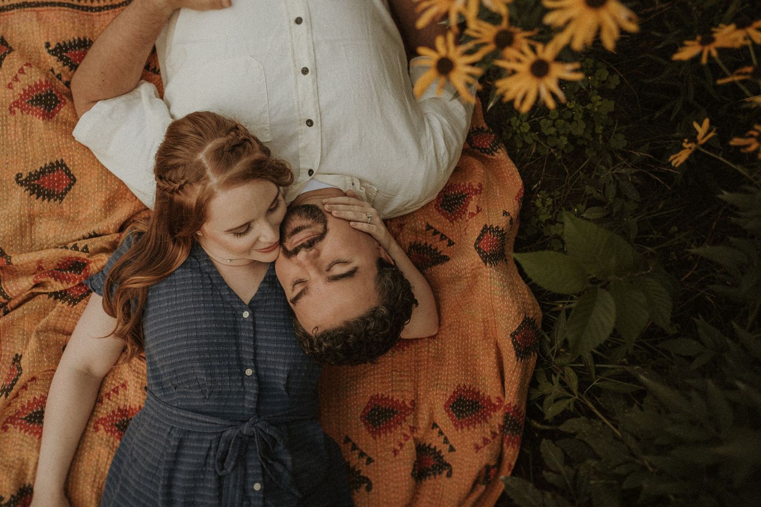 Couple lying on a patterned blanket surrounded by flowers in an outdoor setting.