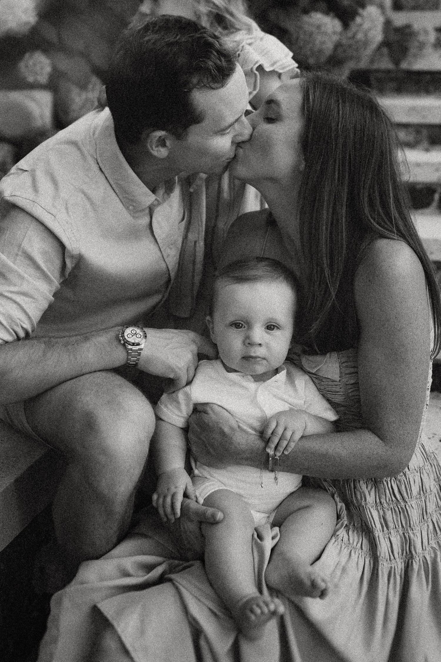 Intimate black and white moment of a couple sharing a kiss while holding their baby on outdoor steps.