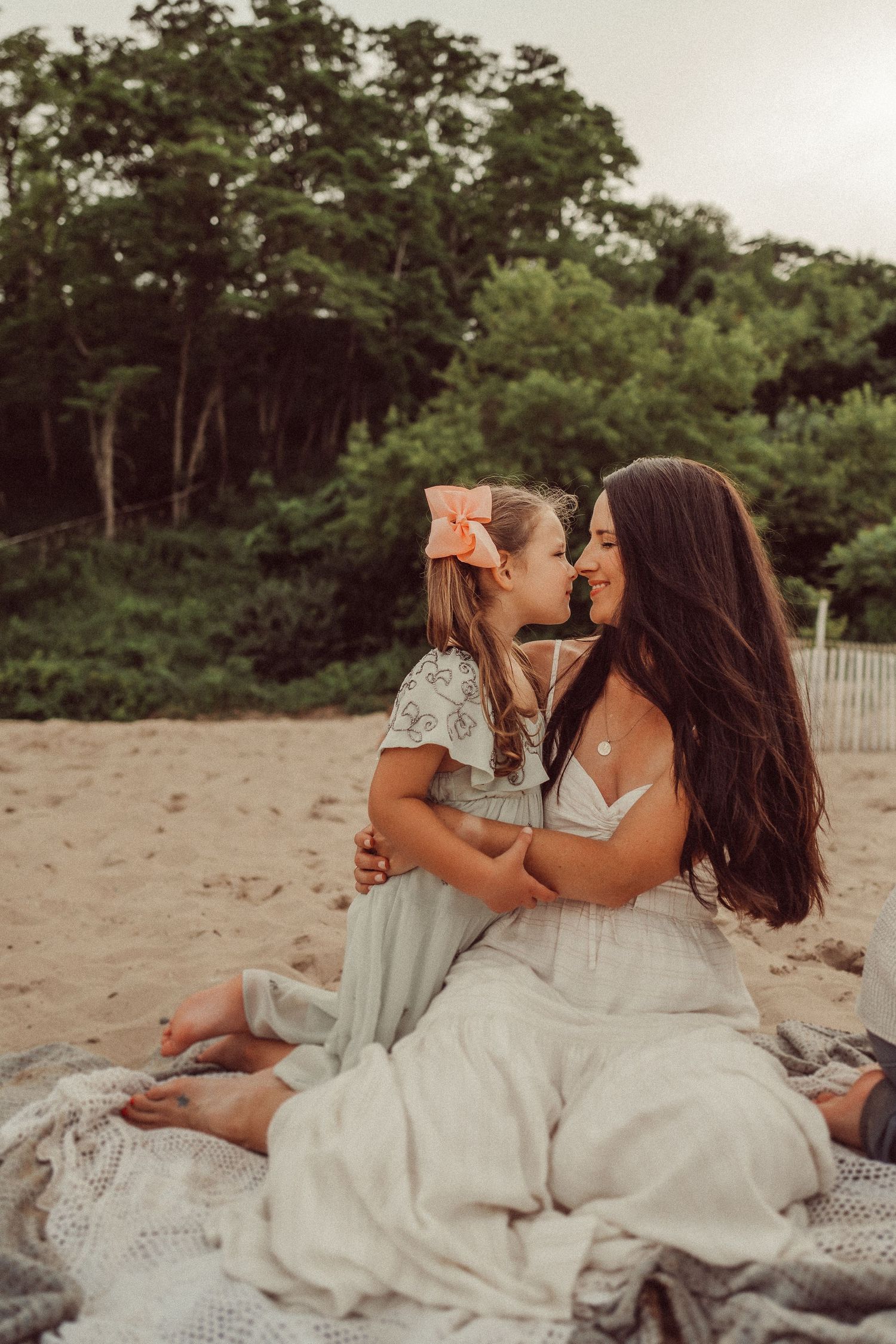 Two people in white dresses share laughter while sitting together on a beach blanket.