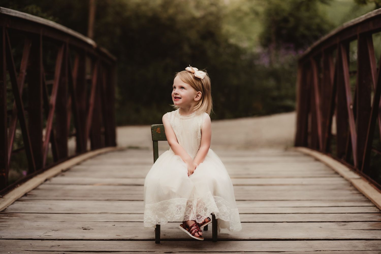Young child in a white dress sitting on a rustic wooden bridge in an outdoor setting.