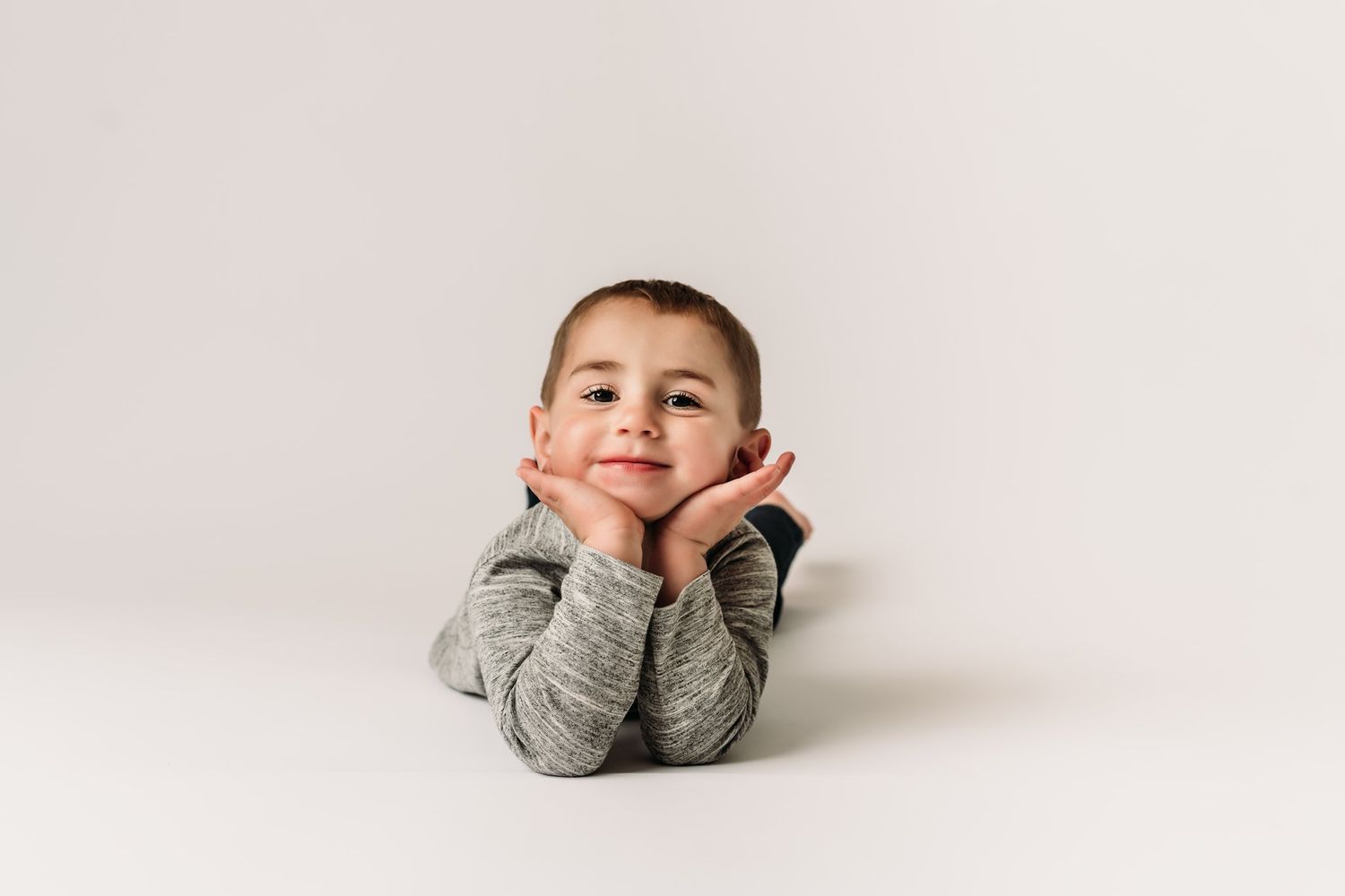 Resting head on hands while lying on stomach on white background wearing gray sweater.