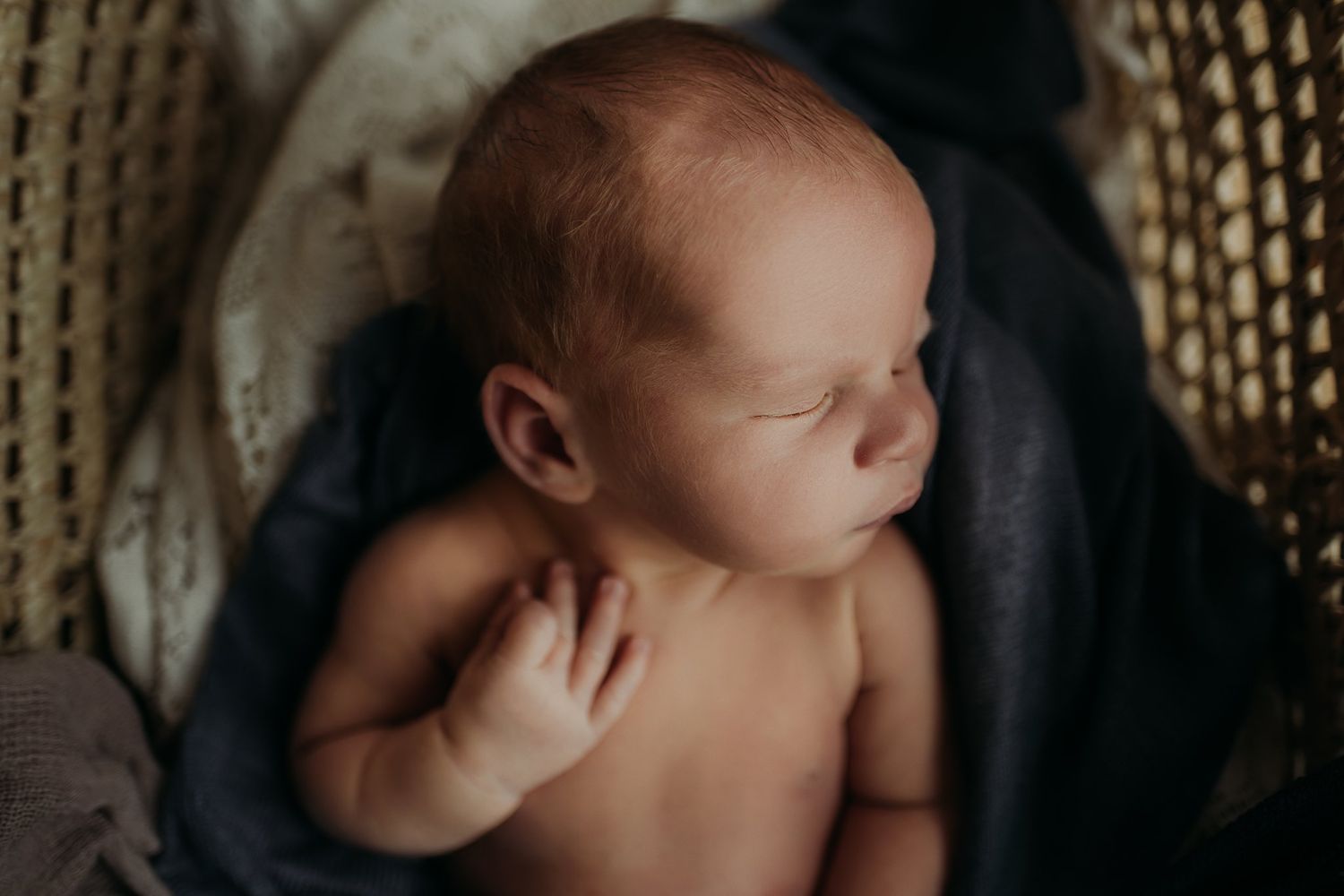 Peaceful newborn sleeps in wicker basket draped with navy blue blanket.