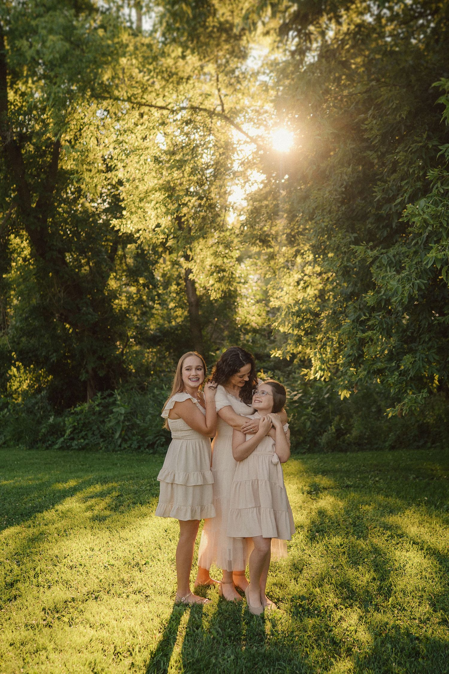 Three friends in coordinated cream dresses embrace while standing in dappled sunlight on a grassy lawn.
