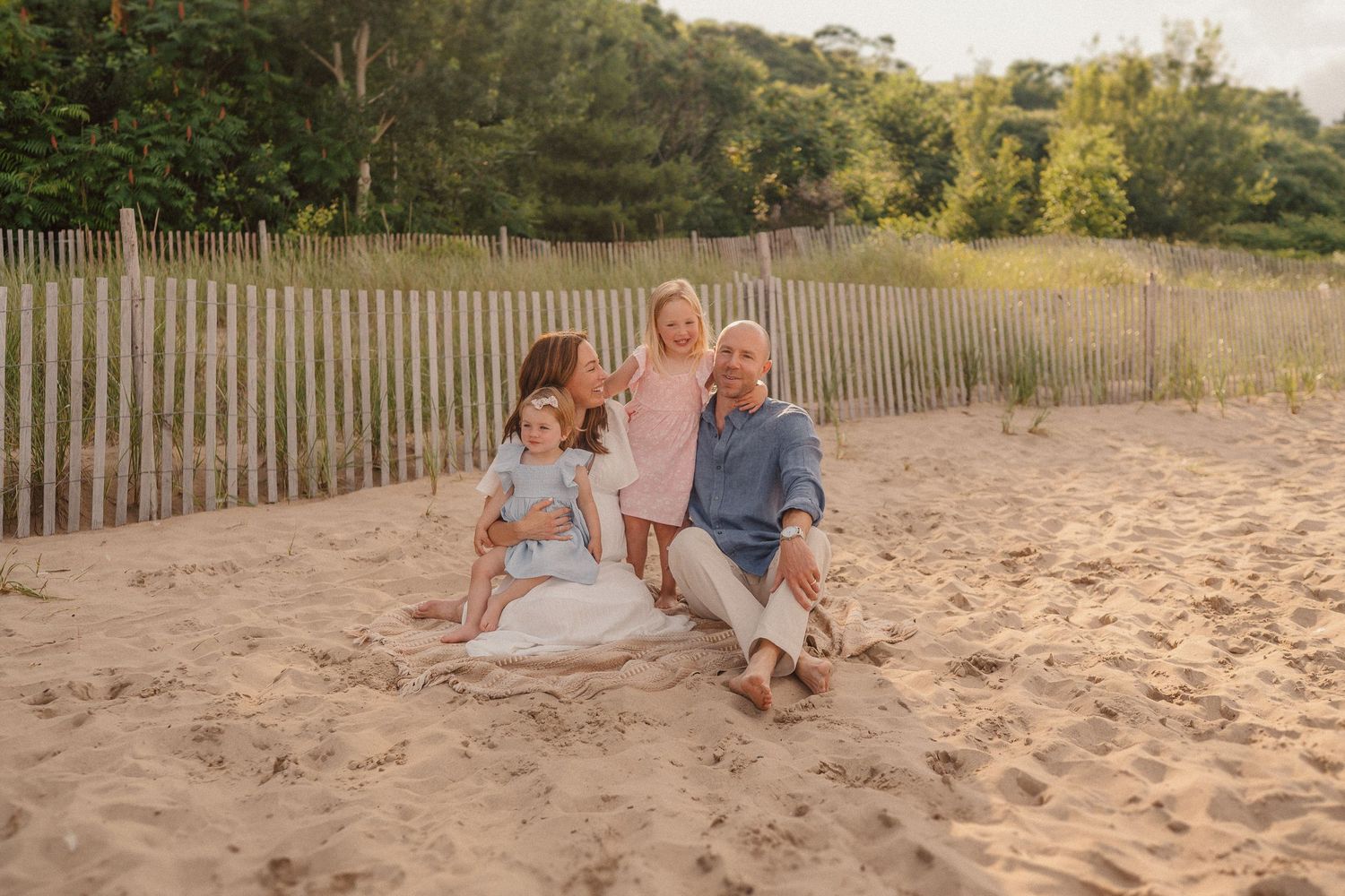 A sunset beach photo with a family group sitting together on sandy shores near wooden fencing and trees.