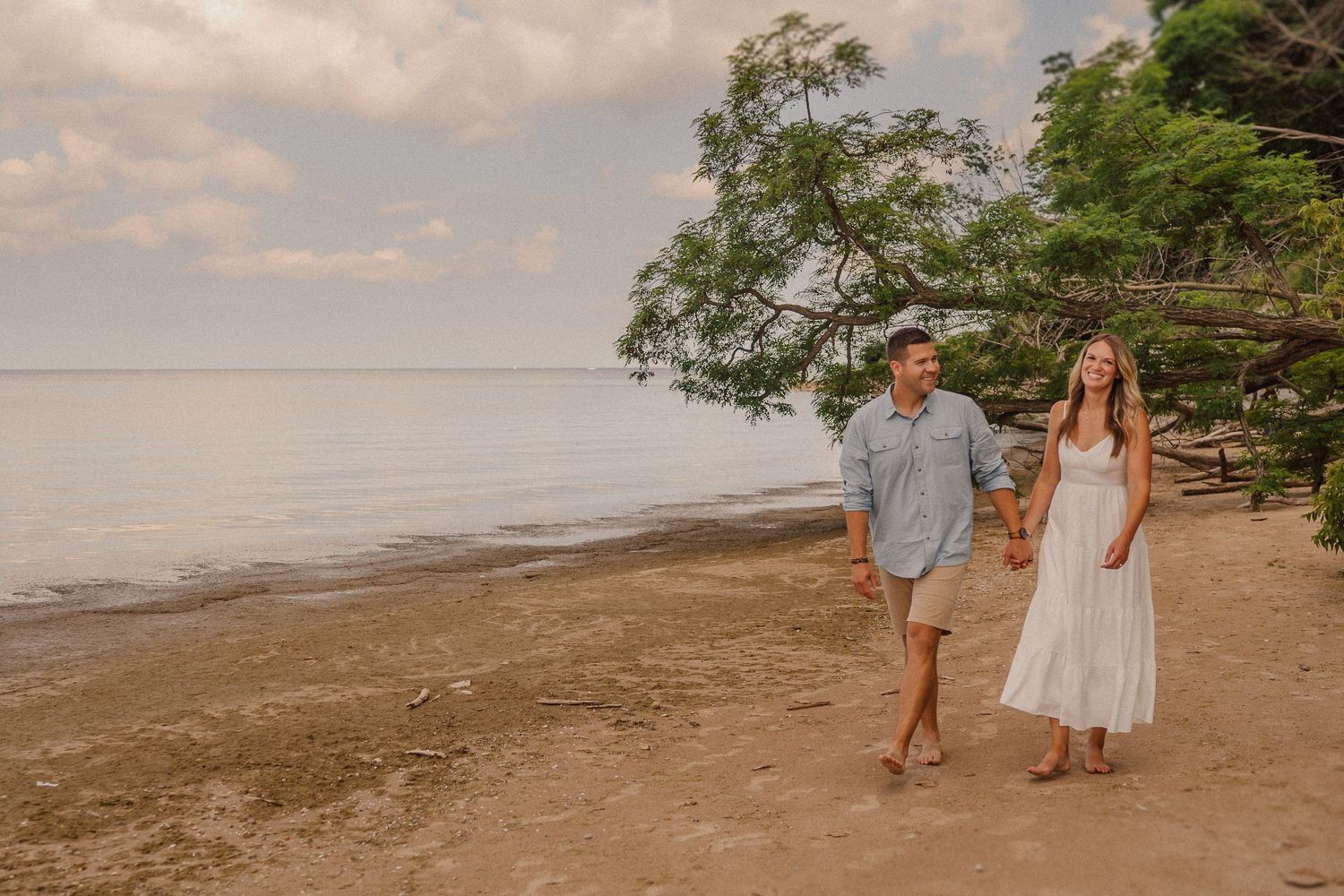 Couple strolling on a peaceful beach with overhanging trees and cloudy skies.