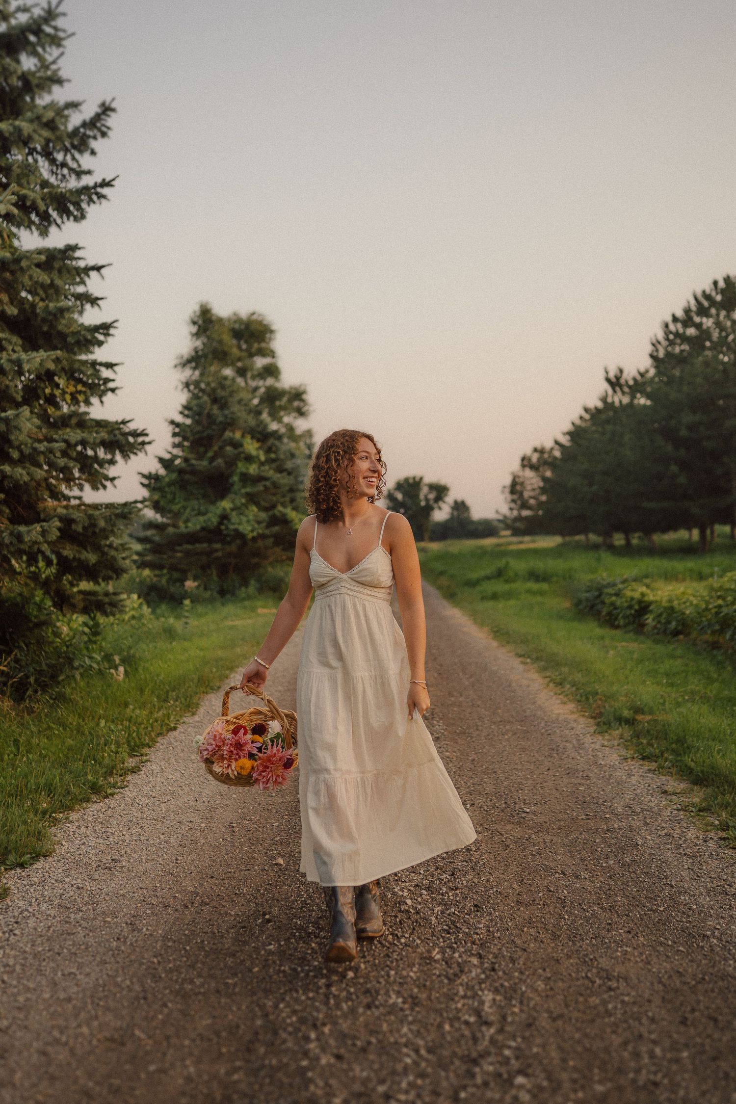 Person in long flowing dress holding flower basket walks along scenic gravel path at golden hour.