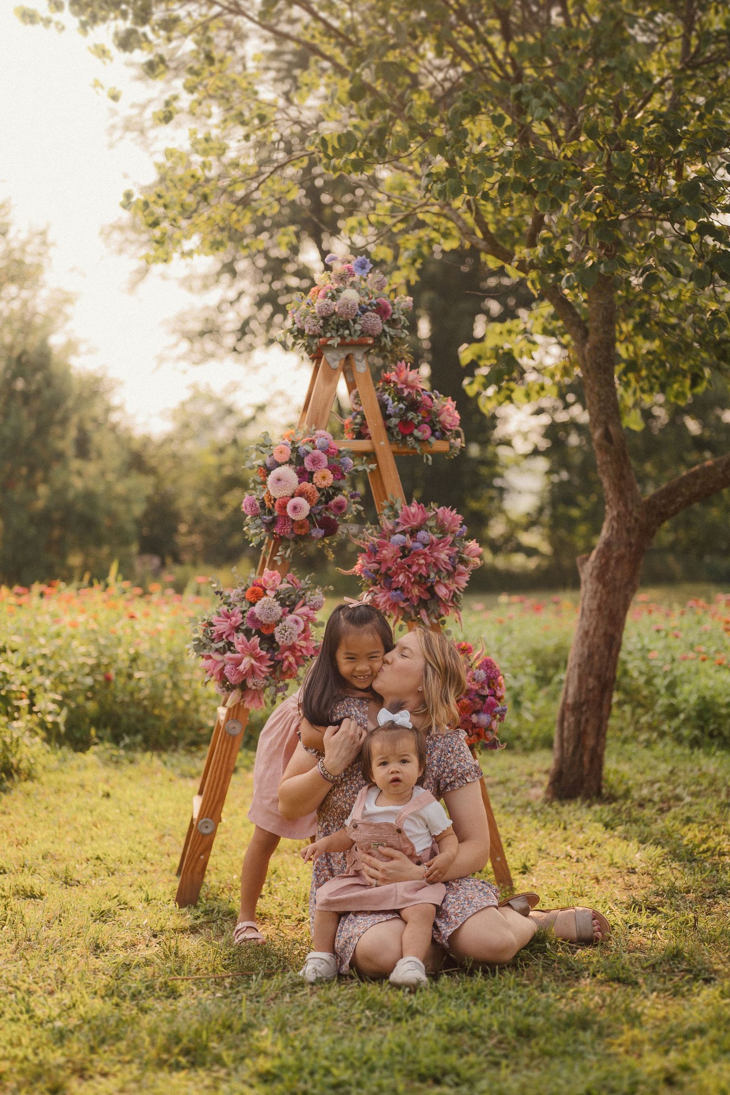Mother and baby sharing tender moments on grass surrounded by pink flowers and sunlit trees.