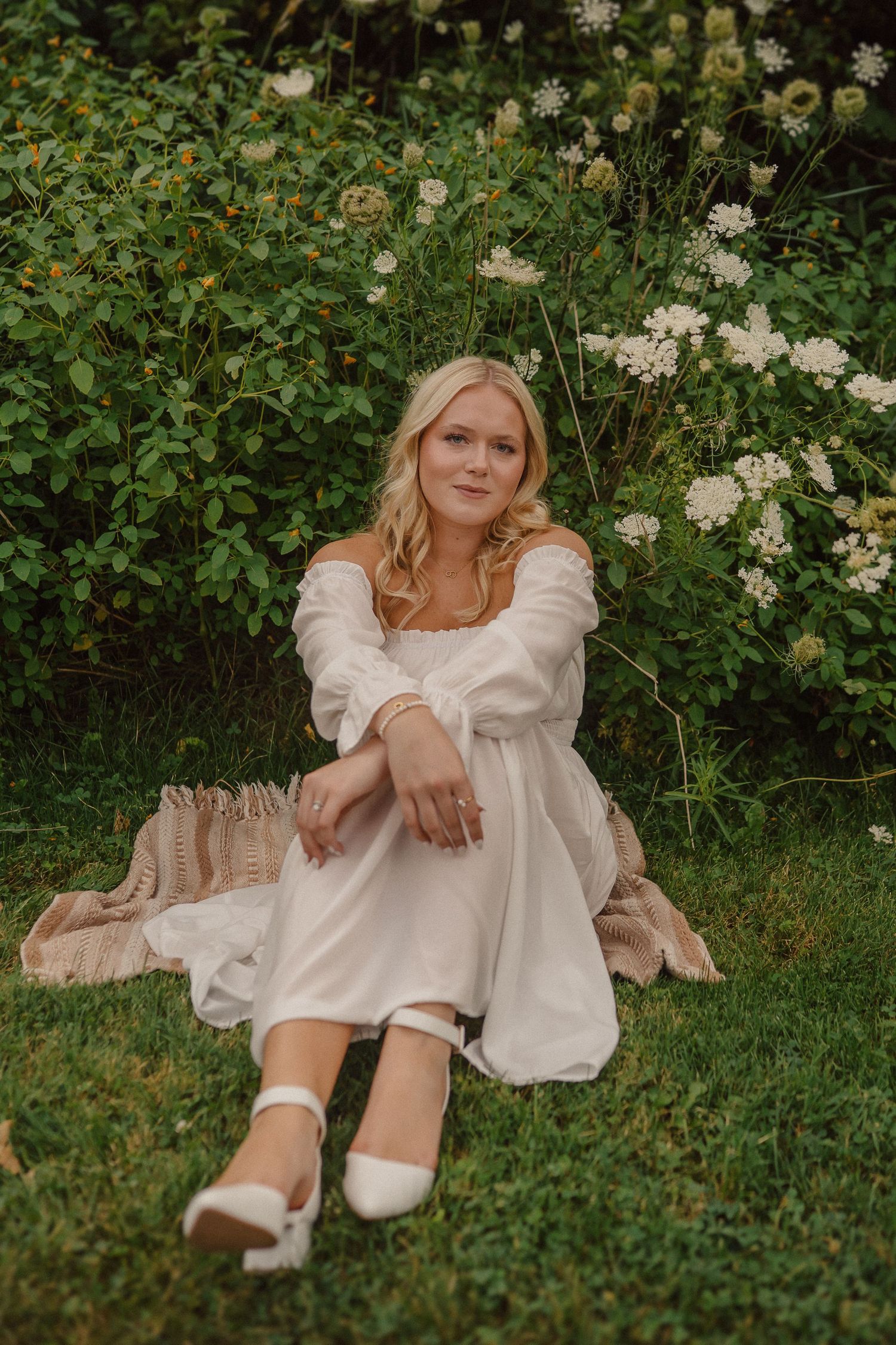 A person in a flowy white dress sits on grass surrounded by white flowers and greenery in a garden setting.