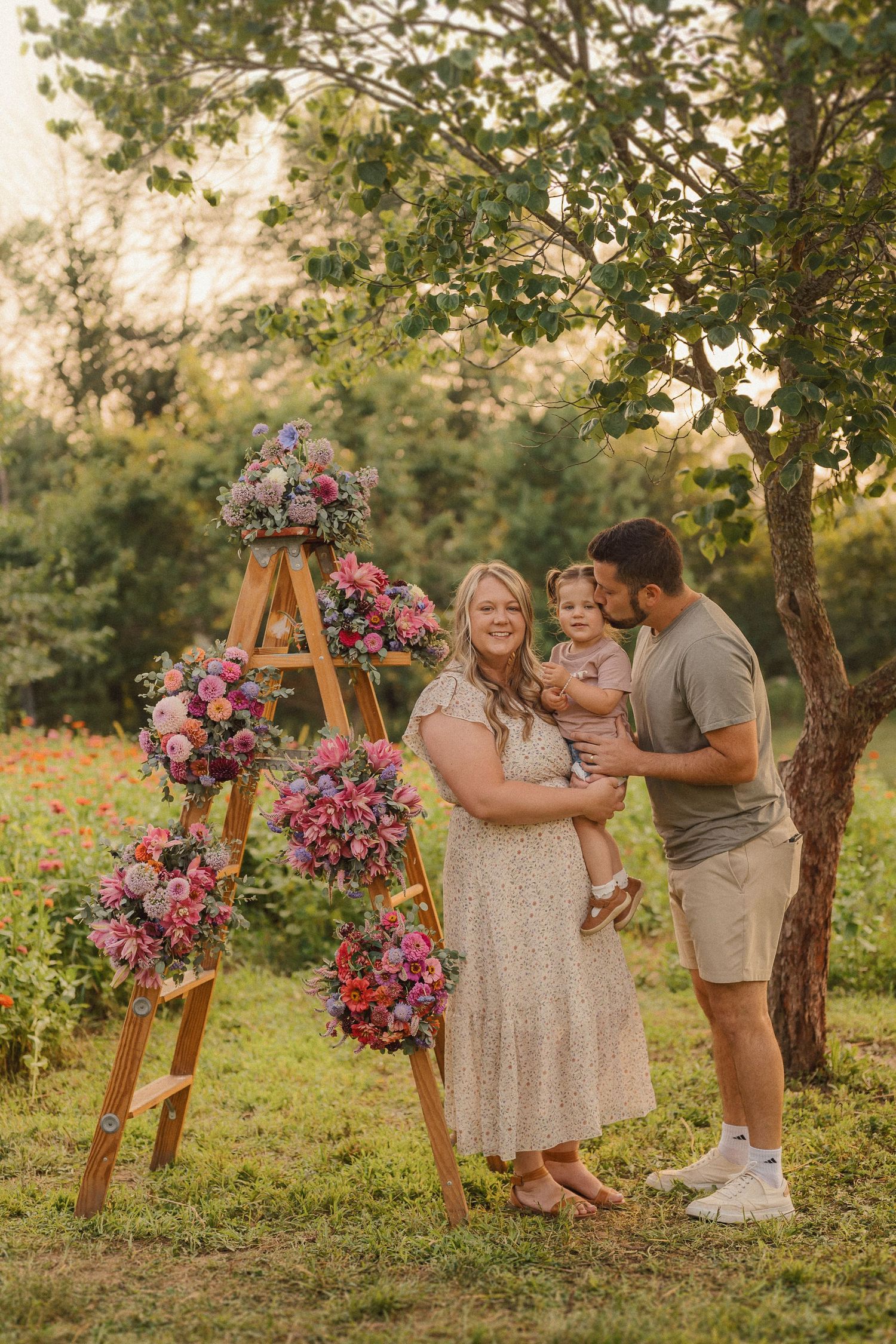 A small family sits together in an outdoor garden setting with wooden floral stands during golden hour.