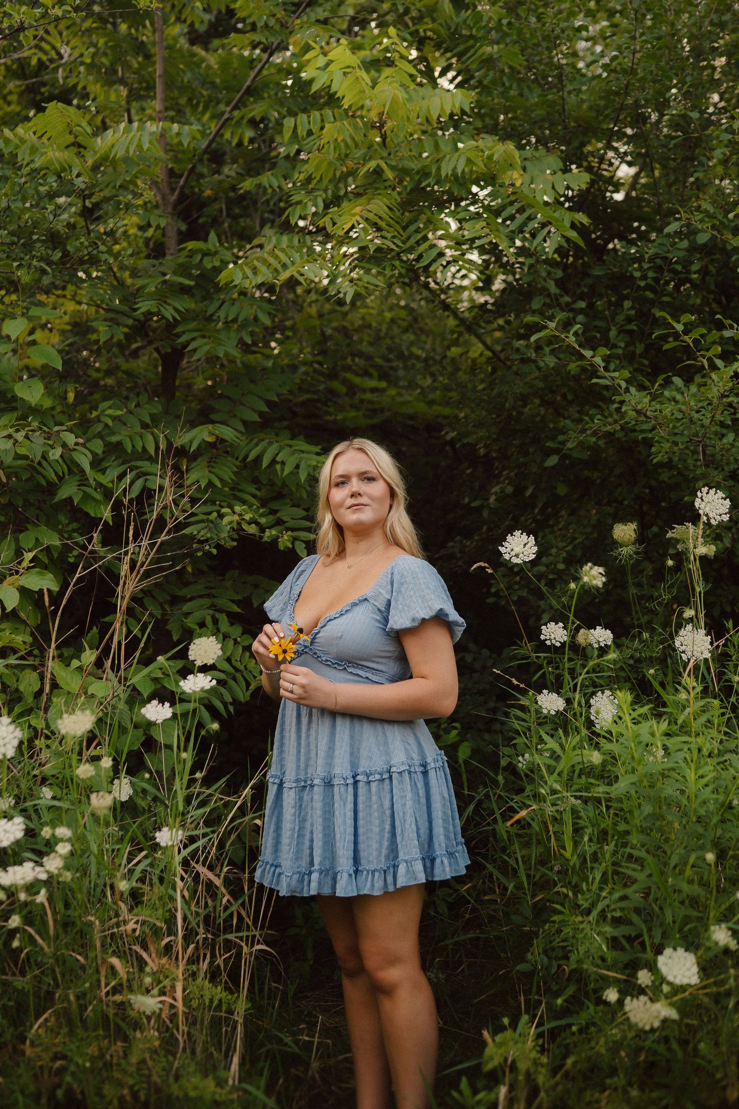 Figure in a light blue babydoll dress poses among blooming white flowers and green foliage.