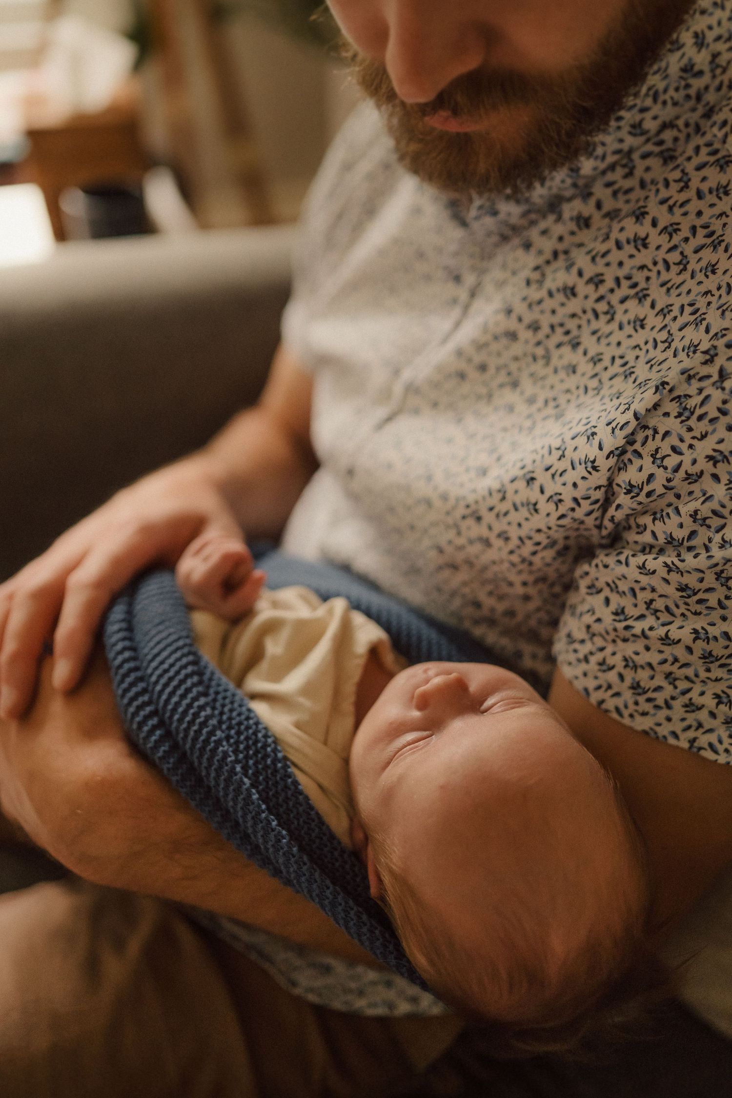 A sleeping baby in a blue knit swaddle is held in caring hands while resting on a cozy sweater.