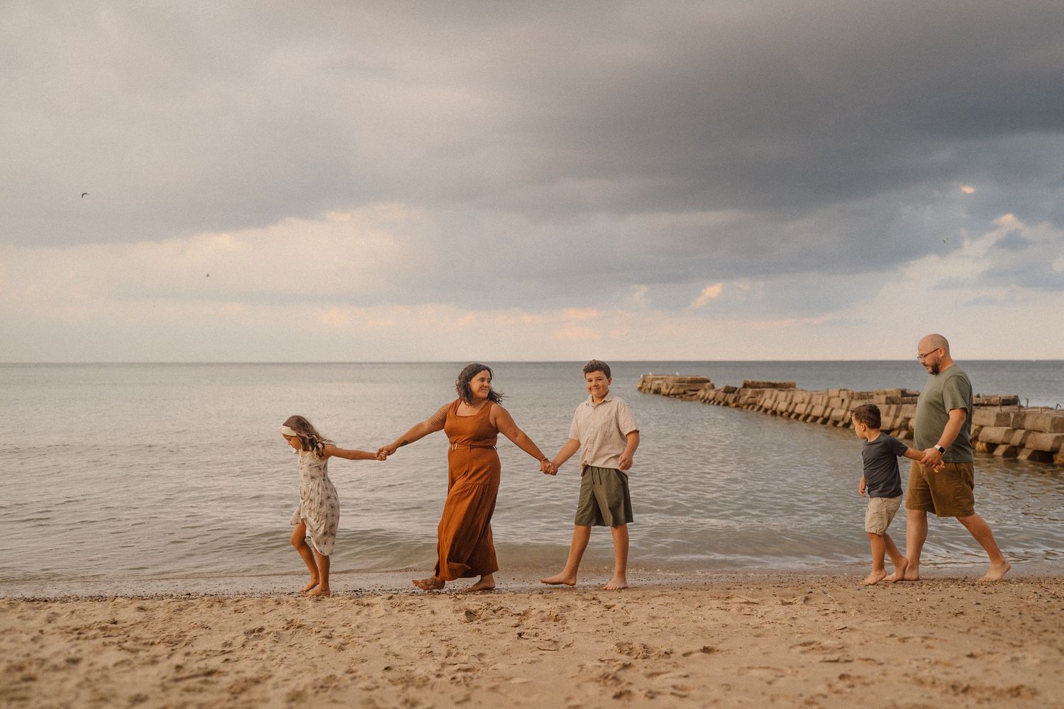 Group of people holding hands and walking along a sandy beach at sunset with a pier in the background.
