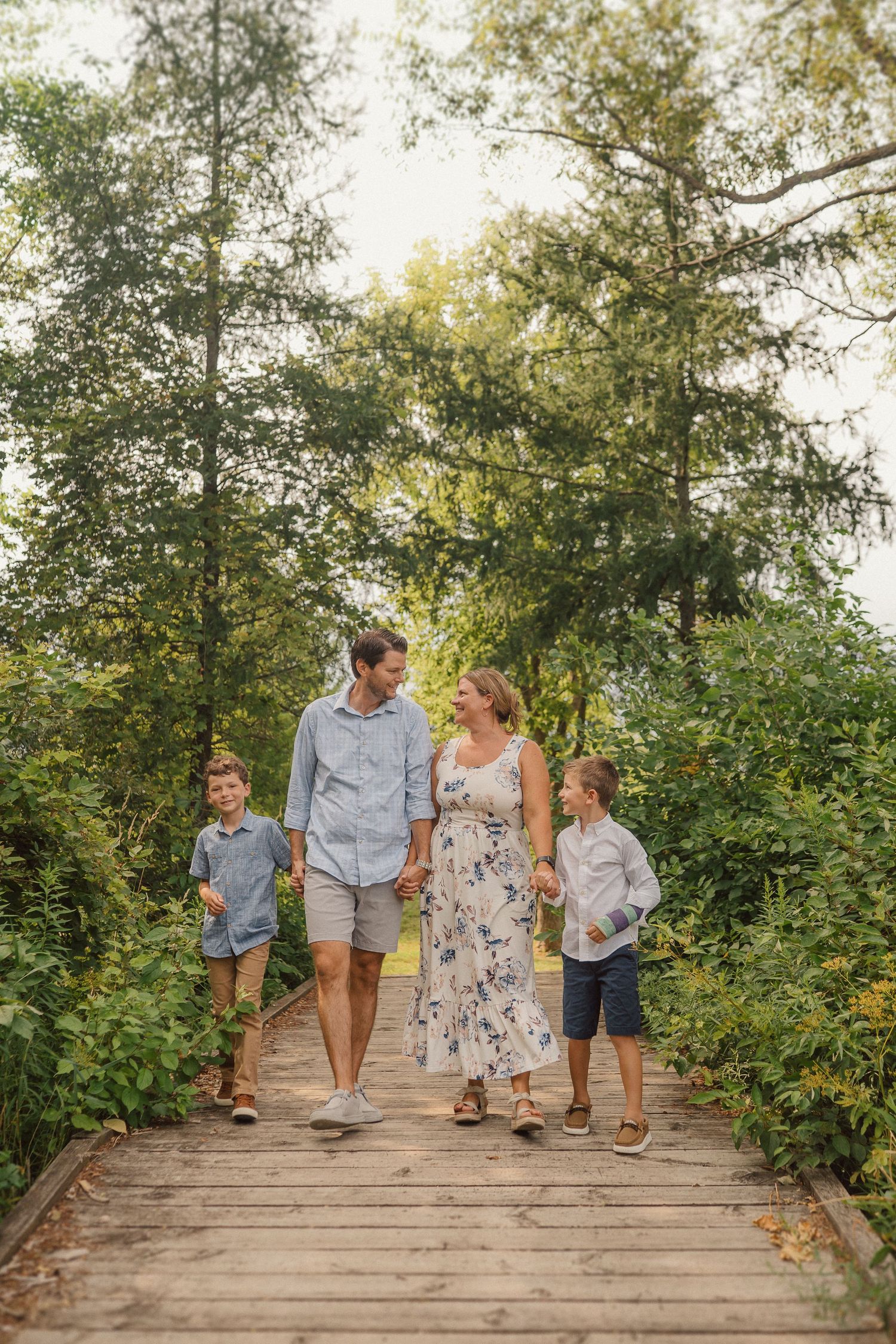 A family strolls together on a wooden boardwalk path through lush green summer foliage.
