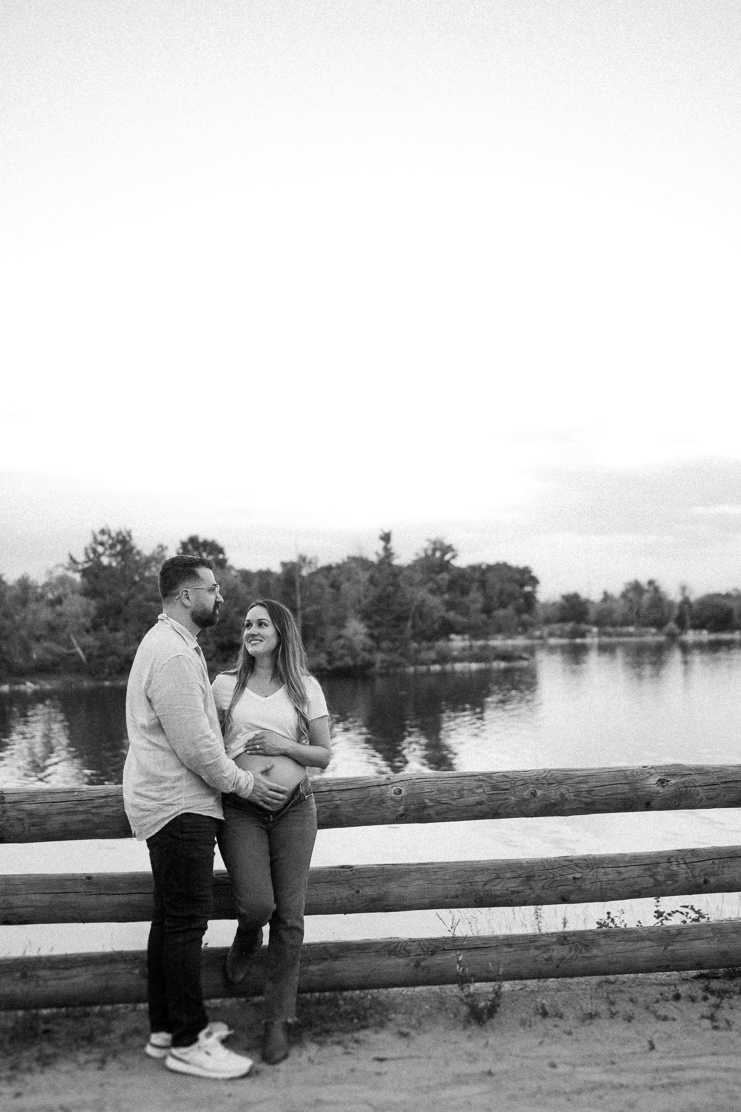 A couple shares a tender moment while leaning against a wooden railing by a lake.