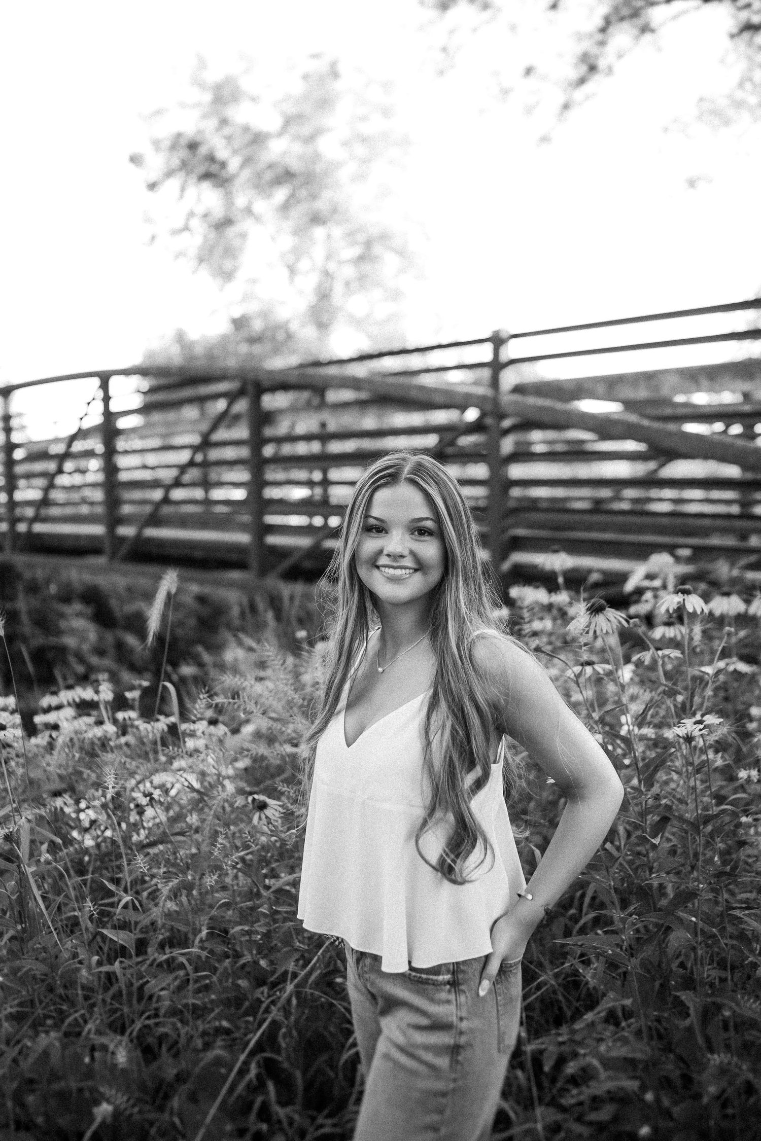 A black and white portrait of someone posing in a flowing tank top near a rustic bridge in a garden setting.