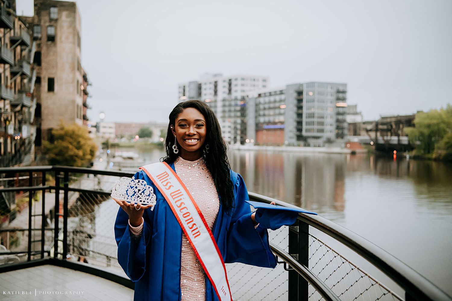 Graduate in blue cap and gown with sash posing on waterfront with buildings in background.