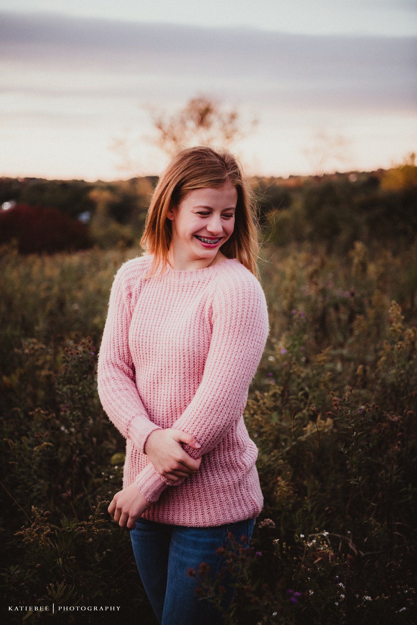 Woman in pink sweater smiling in a field during golden hour sunset.