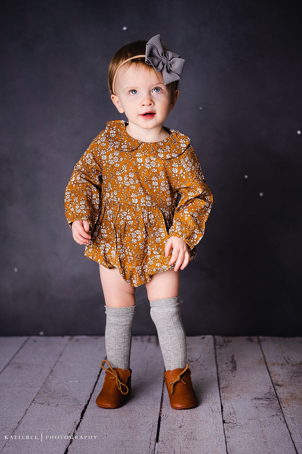 A toddler in knee socks and brown boots wears a mustard floral dress against grey backdrop.