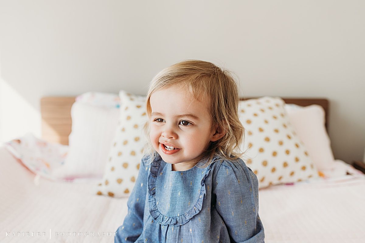 A toddler in blue denim dress smiles while sitting on a bed with gold polka dot pillows.