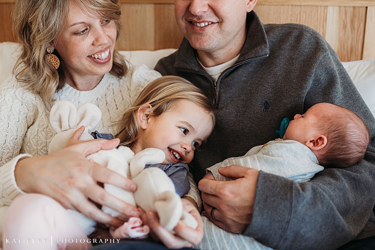 A family of four sits together on a bed sharing a tender moment with their newborn baby.