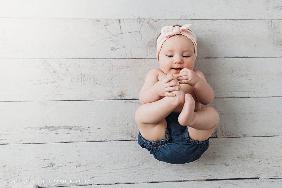 Baby wearing denim and pink headband sits against rustic white wooden wall.