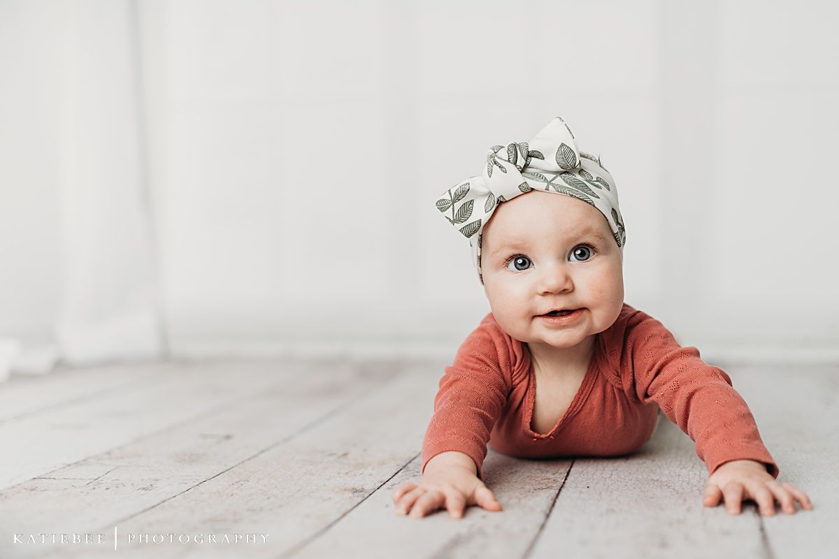 Baby in coral bodysuit and floral headband crawling on white wooden floor.