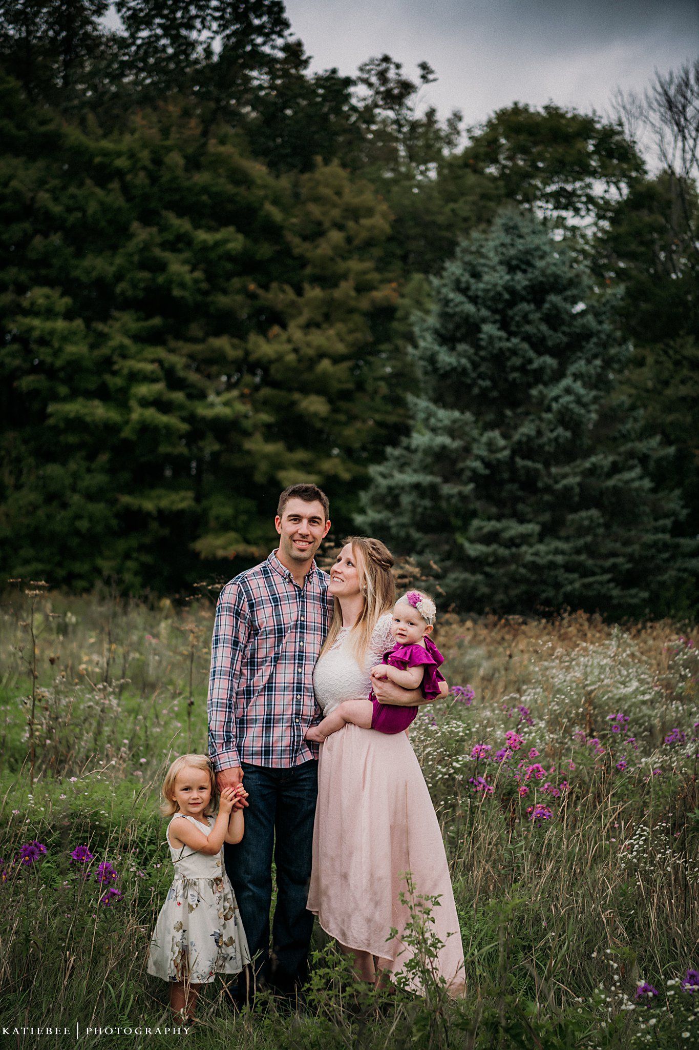 A loving family poses together in a wildflower meadow with evergreen trees in the background.